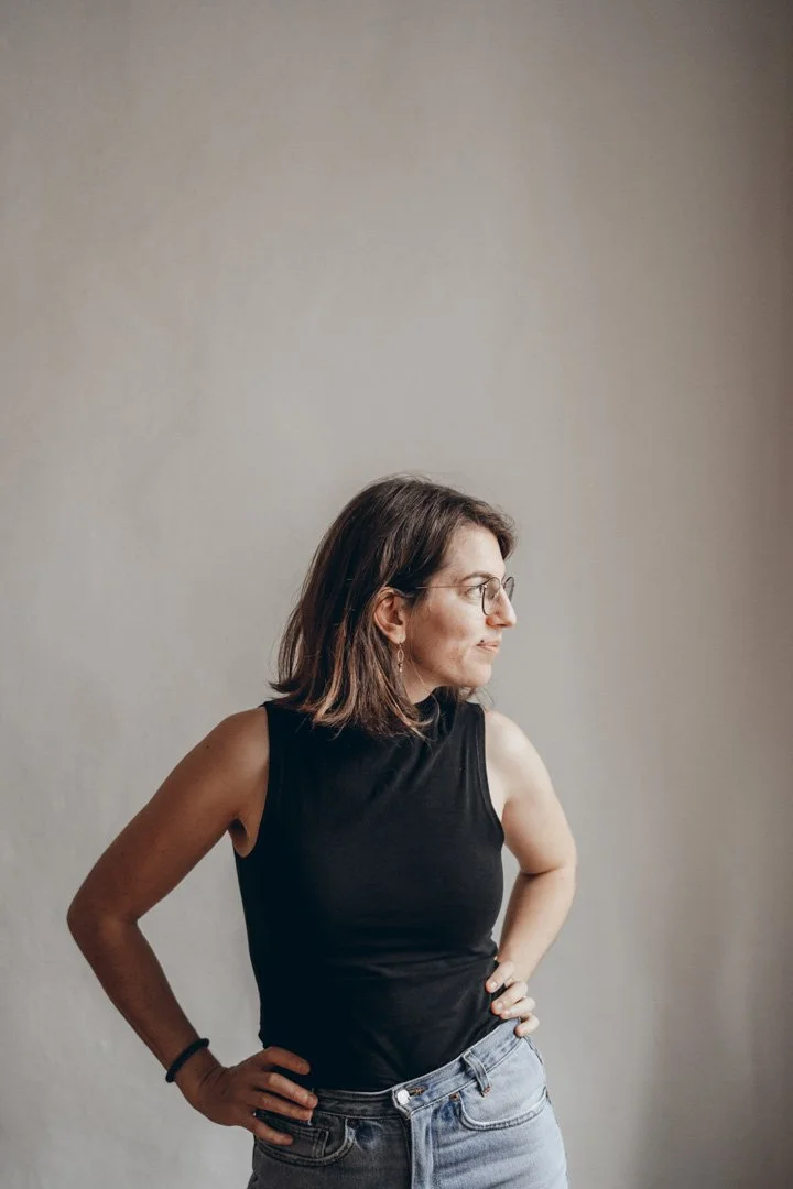 Junge Frau mit Brille in schwarzem Top und Jeans steht seitlich vor einer neutralen Wand.