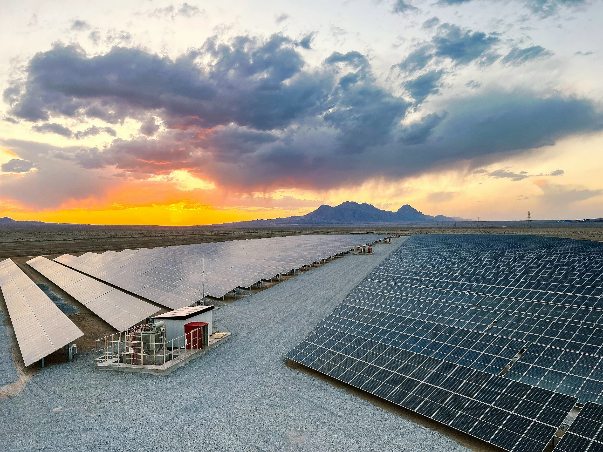 Photovoltaic solar panels installed in an open field during sunset, with mountains and a dramatic sky in the background.