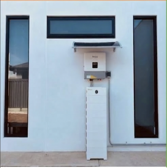 A modern white house exterior with a long, horizontal window and two tall, narrow windows. In front of the house, there is a white electric vehicle charger mounted on the wall, and a white pedestal with a round opening at the bottom, possibly for a trash can or storage. The house has a small overhang with a clear roof above the charger.