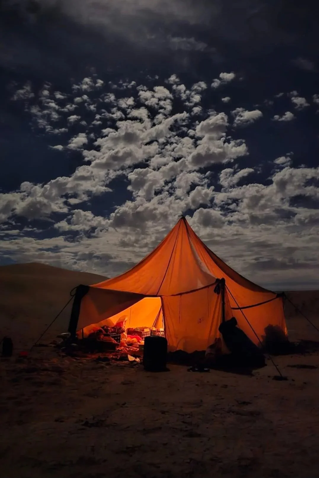Ein orangefarbenes Zelt ist bei Nacht auf einer sandigen Landschaft unter einem bewölkten Himmel mit Sternenhimmel.