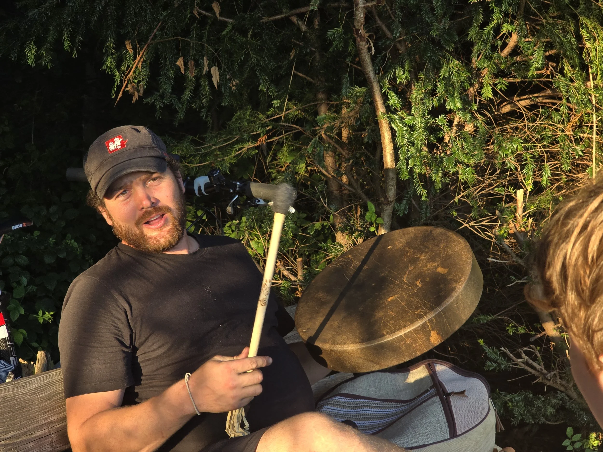 Ein Mann sitzt im Freien vor einem Baum, spielt auf einer Trommel und sieht in die Kamera. Er trägt eine schwarze T-Shirt, eine schwarze Kappe mit einem roten Logo, und hält einen Trommelstock in der Hand.