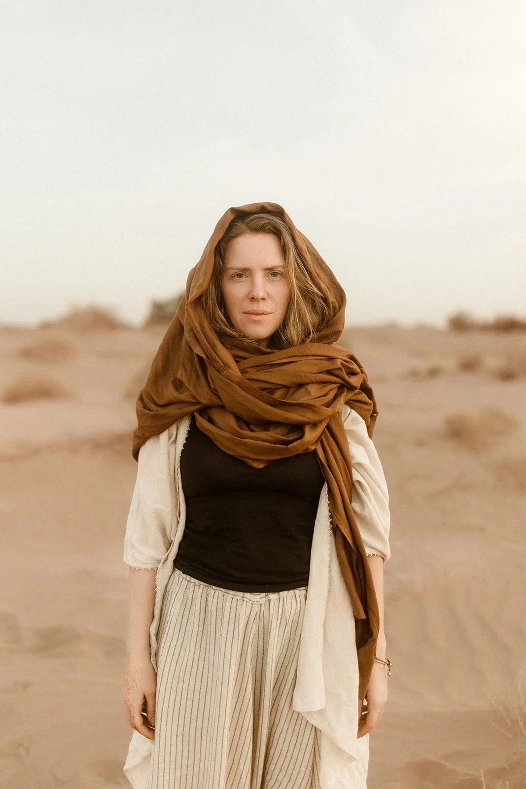 Frau steht in der Wüste, trägt einen braunen Schal über dem Kopf und Schultern, schwarze Top und beige, gestreifte Hosen.