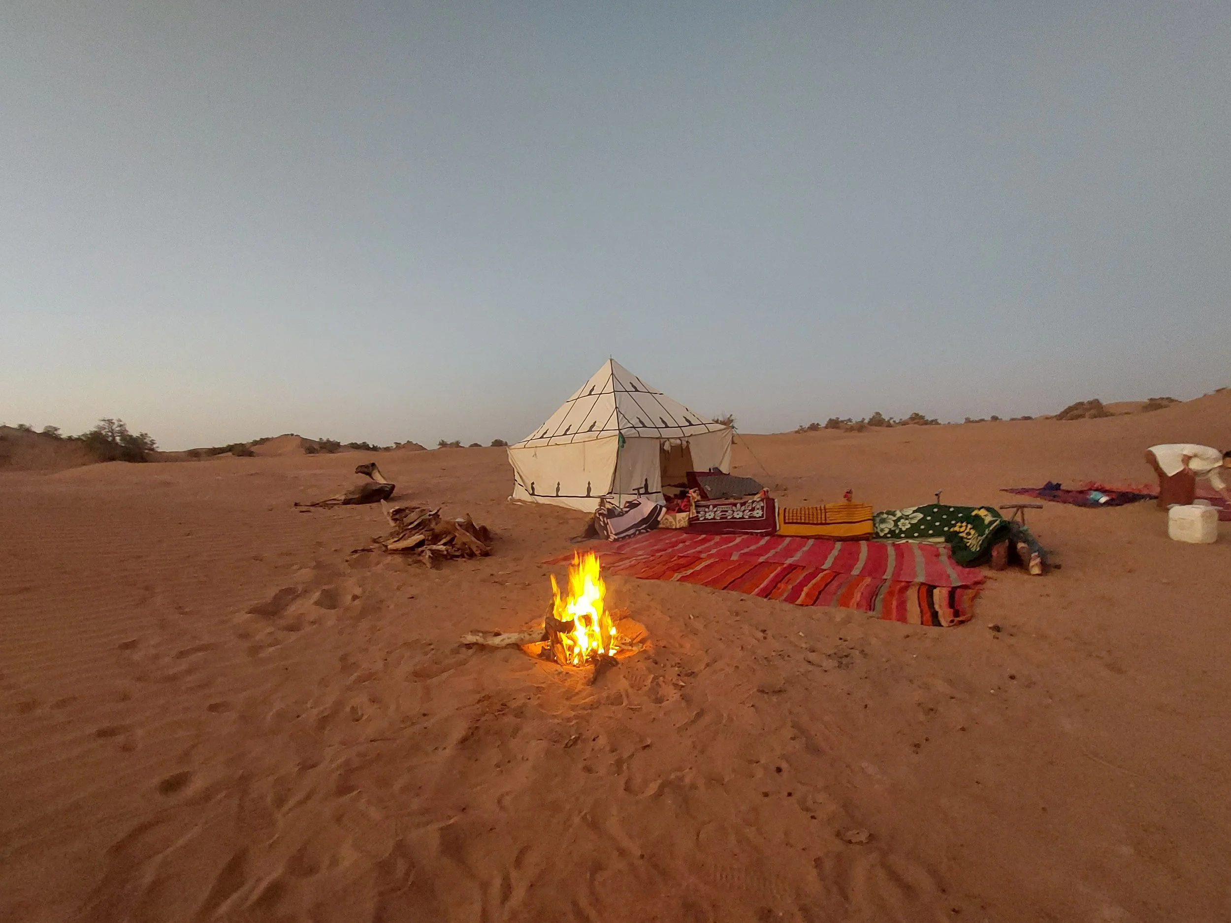 Zelt in einer Wüste bei Sonnenuntergang, umgeben von Teppichen, Kissen und einem Lagerfeuer.