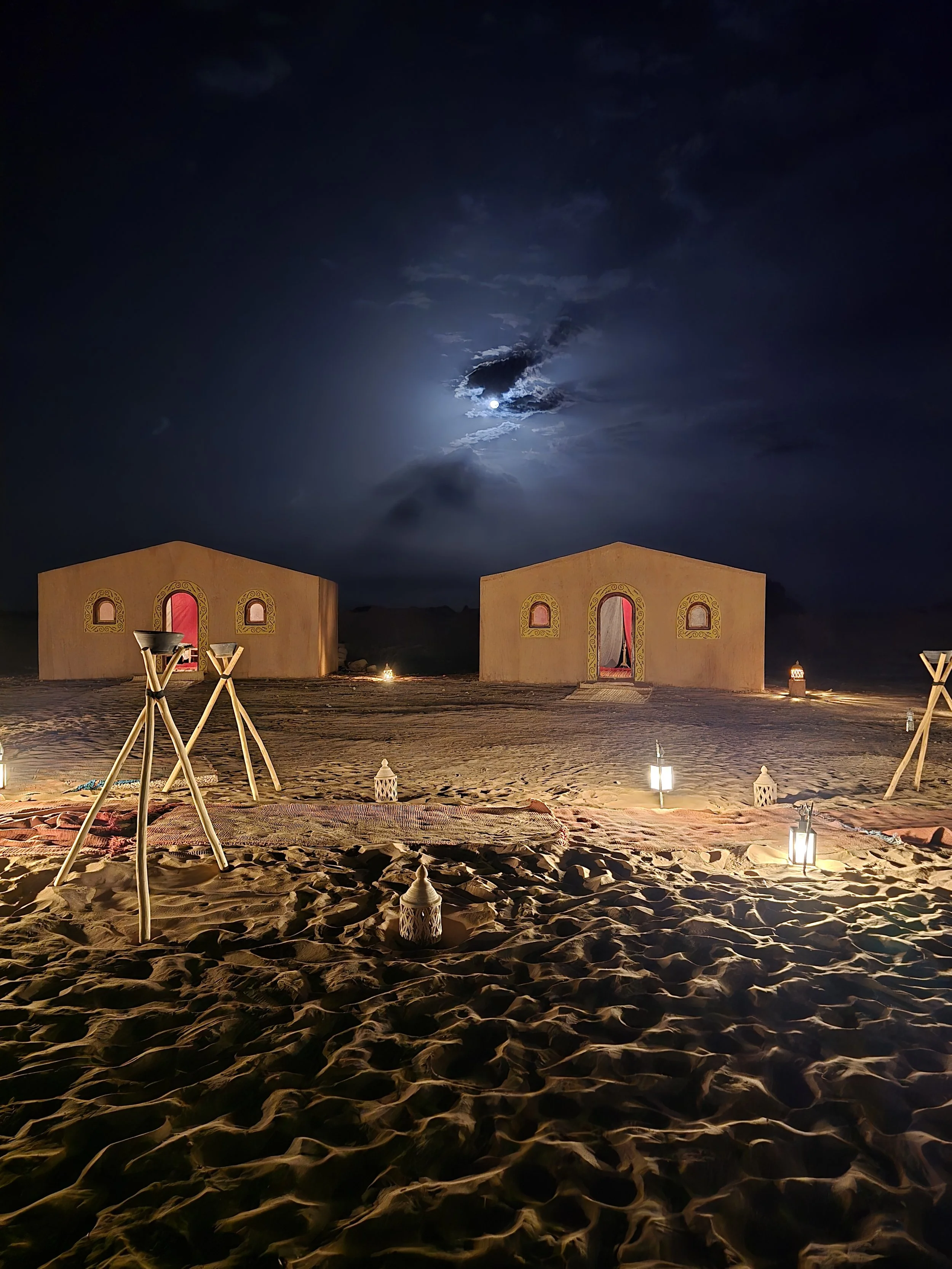 Zwei Zelte in der Wüste bei Nacht, beleuchtet von Laternen, mit dem Mond und Wolken am Himmel im Hintergrund.