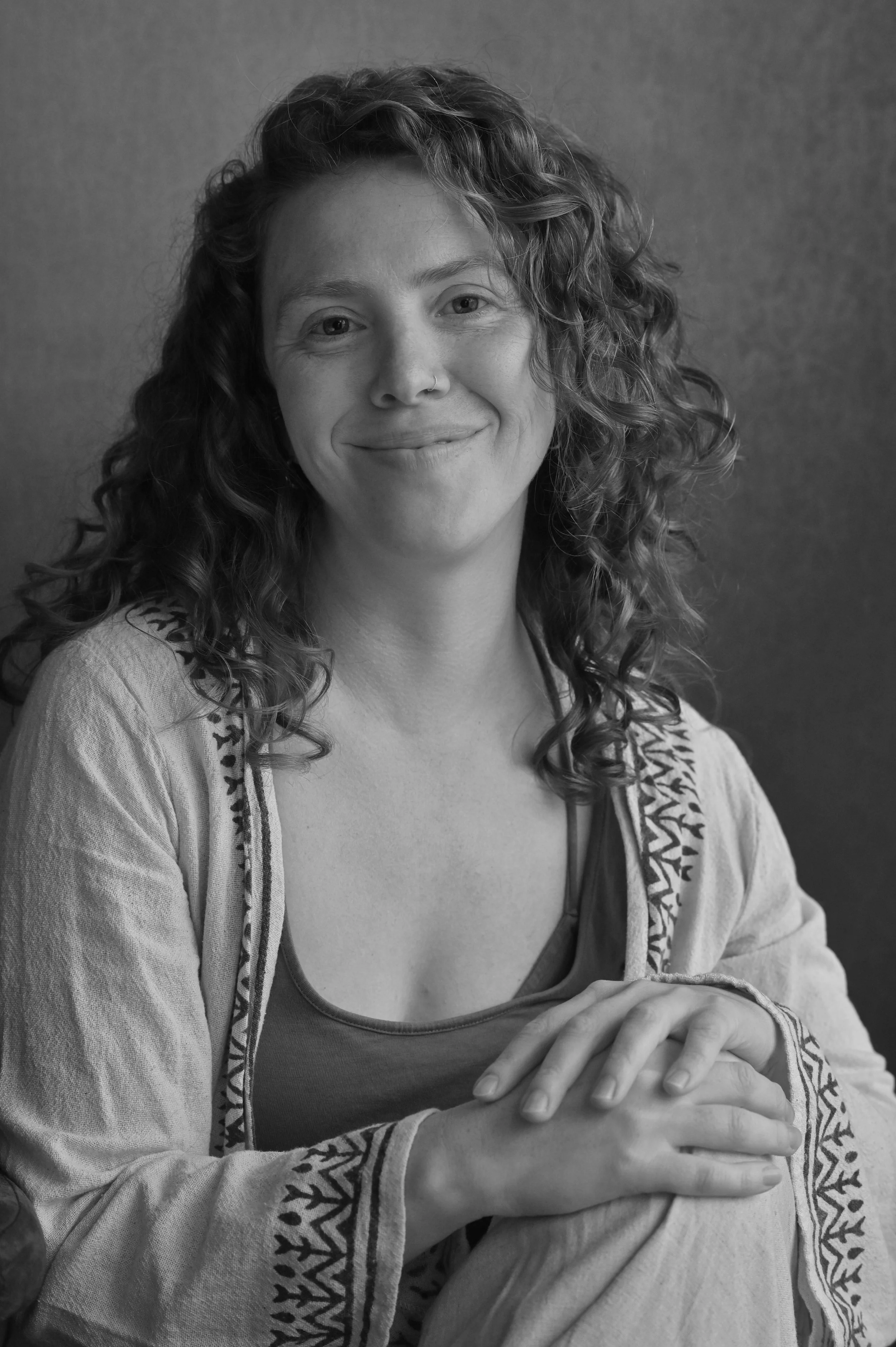 Schwarz-Weiß-Porträt einer lachenden Frau mit lockigem Haar, die auf einer ländlichen Bank sitzt. Sie trägt ein süßes top und eine Jacke mit ethnischem Muster.