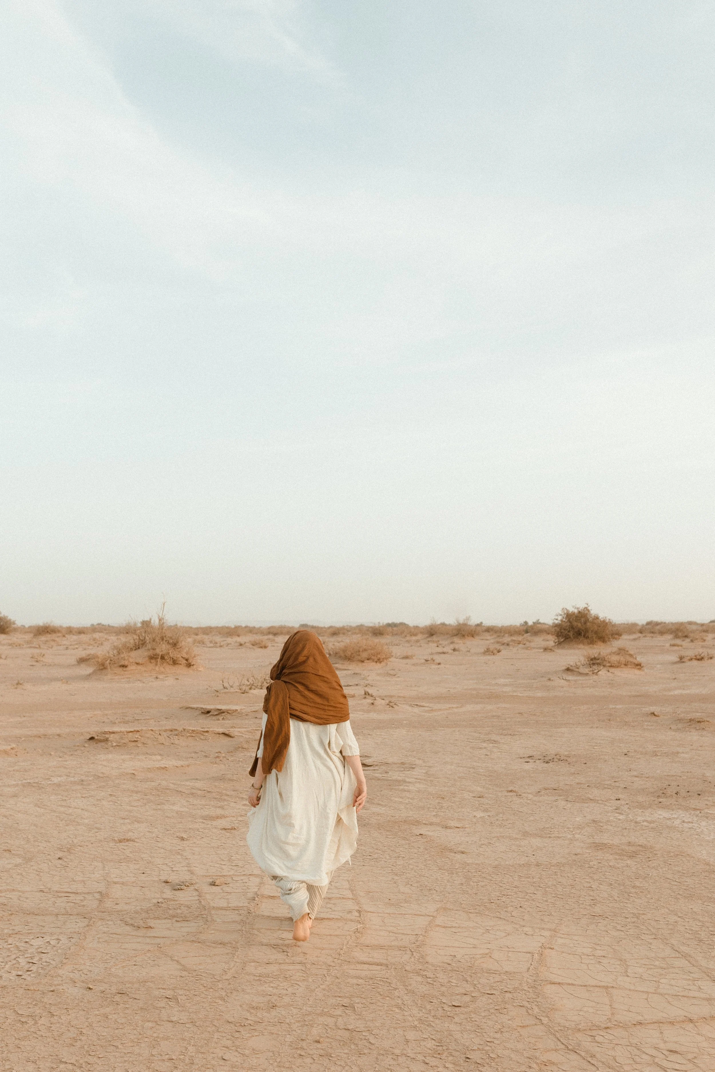 Eine Person, die in einem weißen Gewand und einem braunen Kopftuch durch eine trockene, wüstenartige Landschaft läuft.