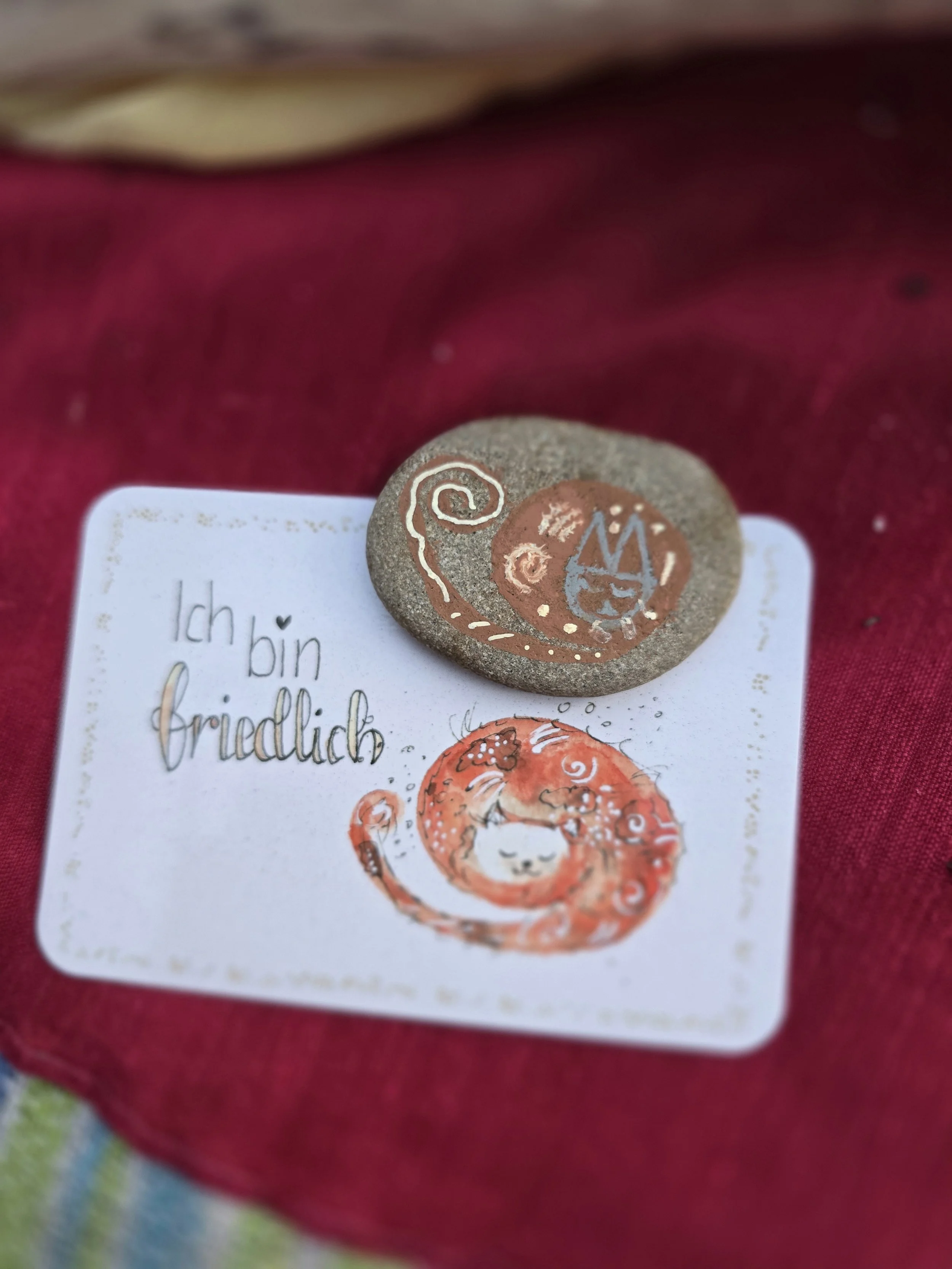Ein kleiner Stein mit einem handgezeichneten Katzenkopf und einer Spirale. Darunter liegt eine Karte mit der Aufschrift "Ich bin friedlich" und einer Zeichnung einer schlafenden Katze in einer Spirale, alles auf einem roten Stoff.