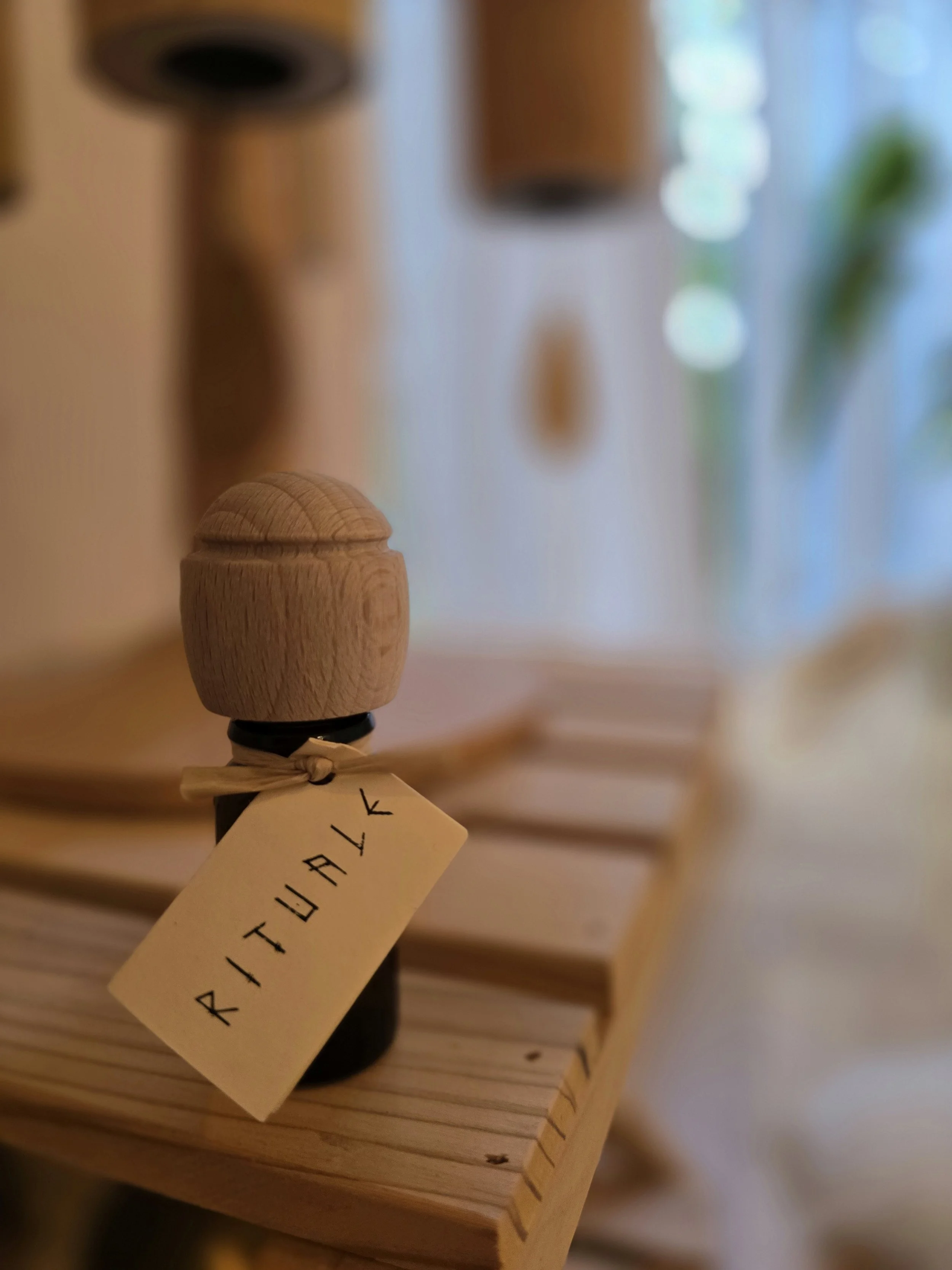 Holzfigur mit einem Tag, auf dem steht 'ATTUNALK', steht auf einem Holzbrett, im Hintergrund unscharfe Innenraumdekorationen.