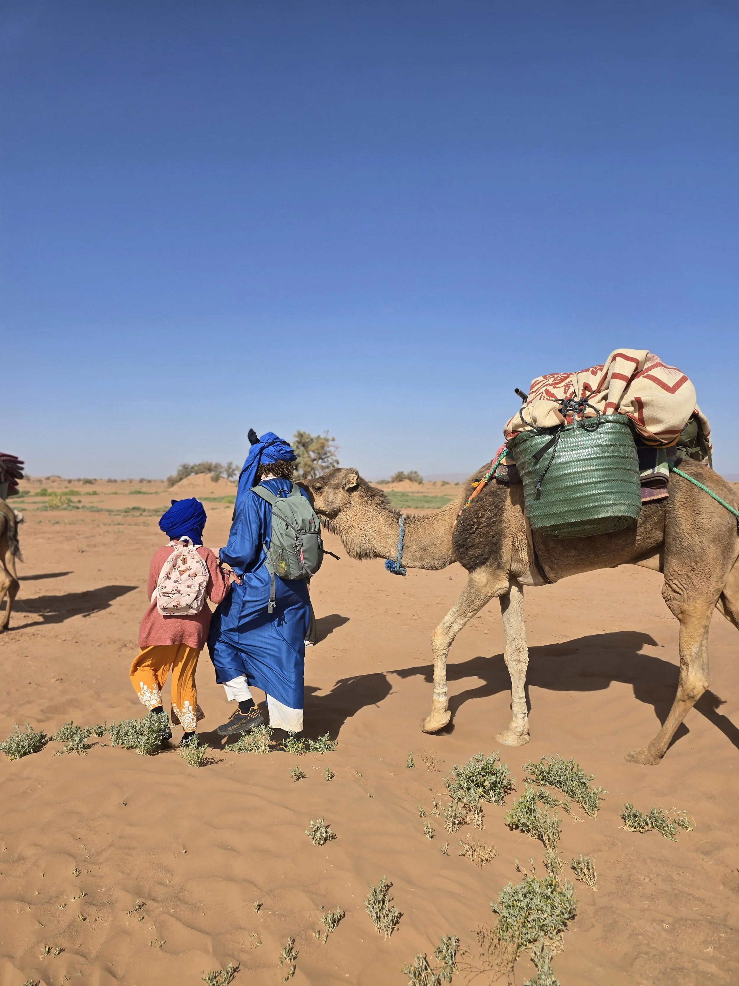 Zwei Personen, ein Mann und ein Kind, mit traditionellen marokkanischen Kleidung, stehen in der Wüste und berühren die Kuppel eines Kamels. Das Kamel ist beladen mit Gepäck und liegt auf dem Sand. Der Himmel ist klar und blau.