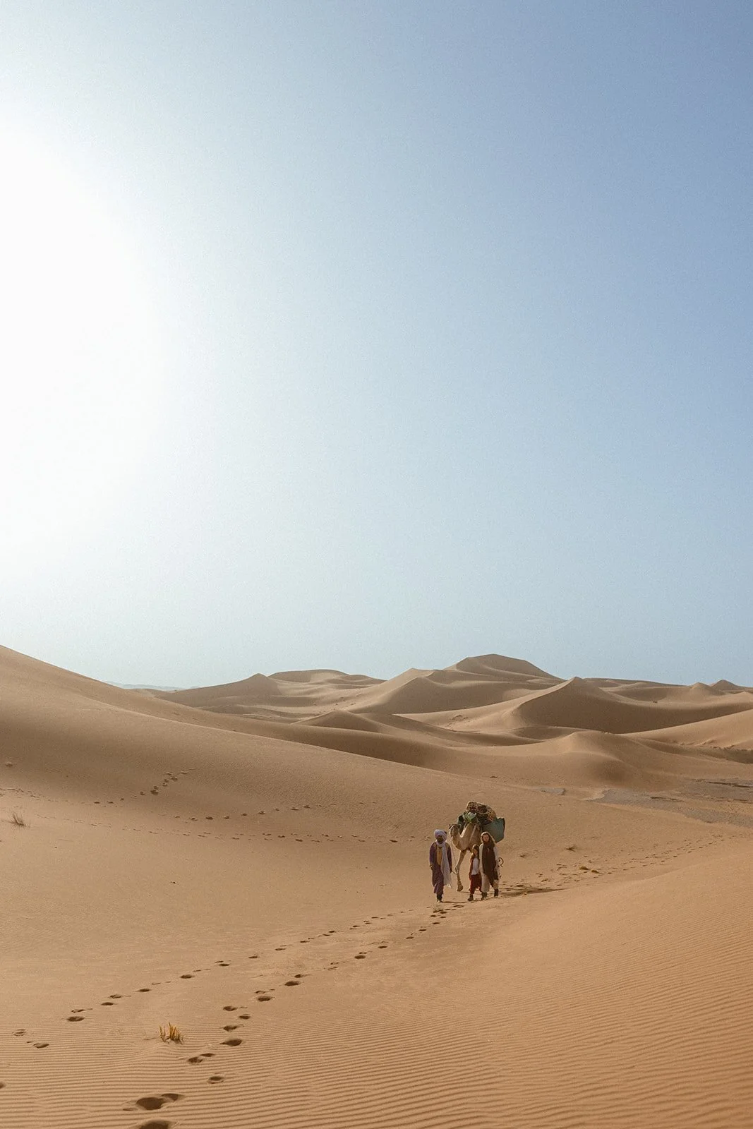 Menschen wandern durch die Wüste mit Kamelen, umgeben von Sanddünen unter einem klaren blauen Himmel.