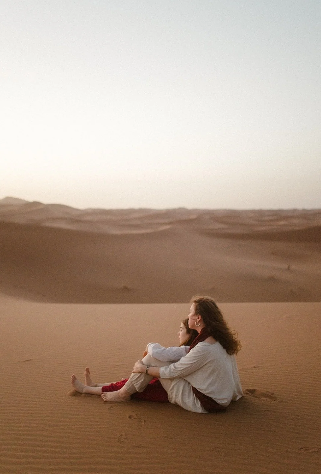 Zwei Frauen sitzen in der Wüste auf Sanddünen, bei Sonnenuntergang, entspannt und in Gedanken versunken.