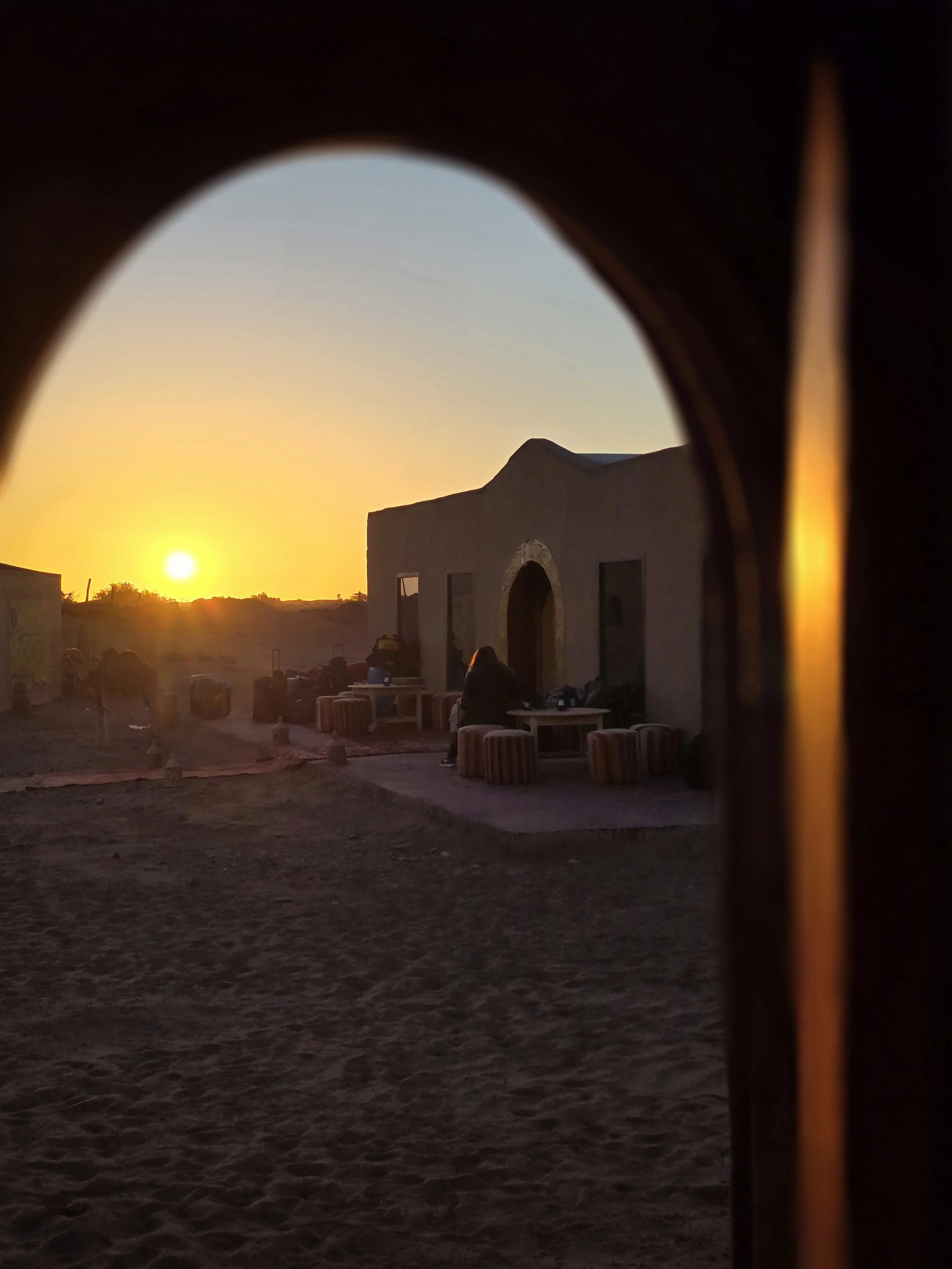 Sonnenuntergang hinter einer weißen, ungewöhnlich geformten Haus- oder Hütte im nordafrikanischen Stil, gesehen durch eine kleine Fensteröffnung in einer dunklen Wand, mit Sitzgelegenheiten und einer Person im Schatten davor.