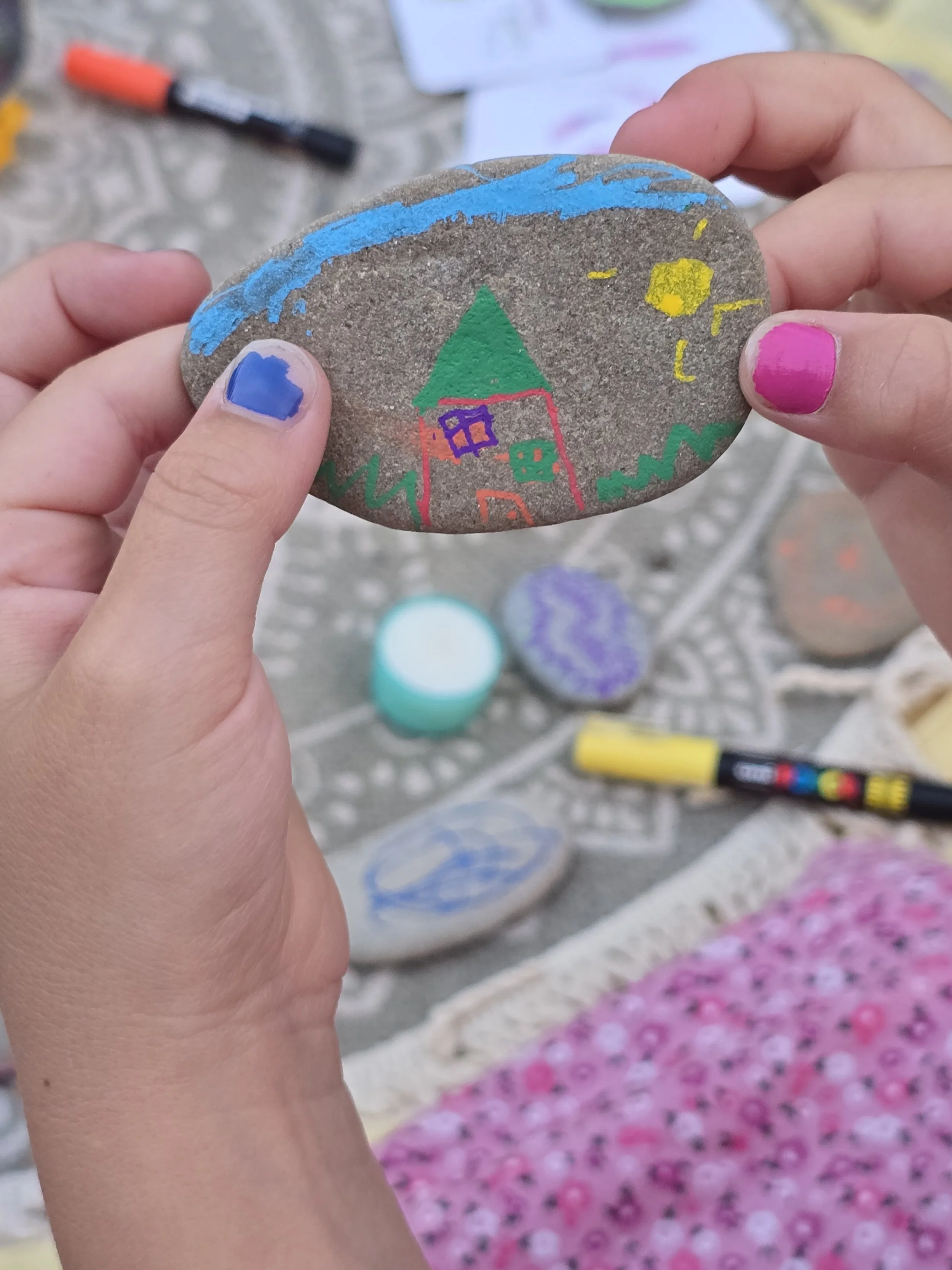 Bemalte Steine, die ein Haus mit grünem Dach, blauen Wasser, gelbem Stern und grünen Bäumen darstellen, mit weiteren bunten Steinen und Markern im Hintergrund.