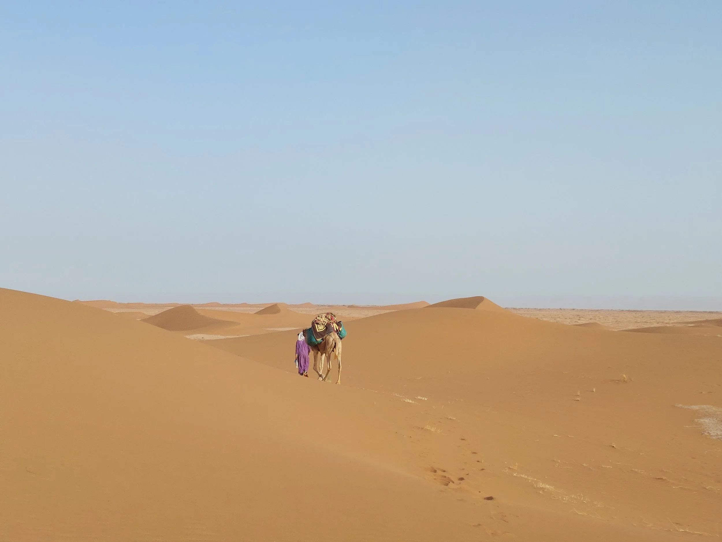 Wüstenlandschaft mit Sanddünen und Himmel, eine Person mit einem Kamel, das mit Gepäck beladen ist, läuft entlang der Düne.