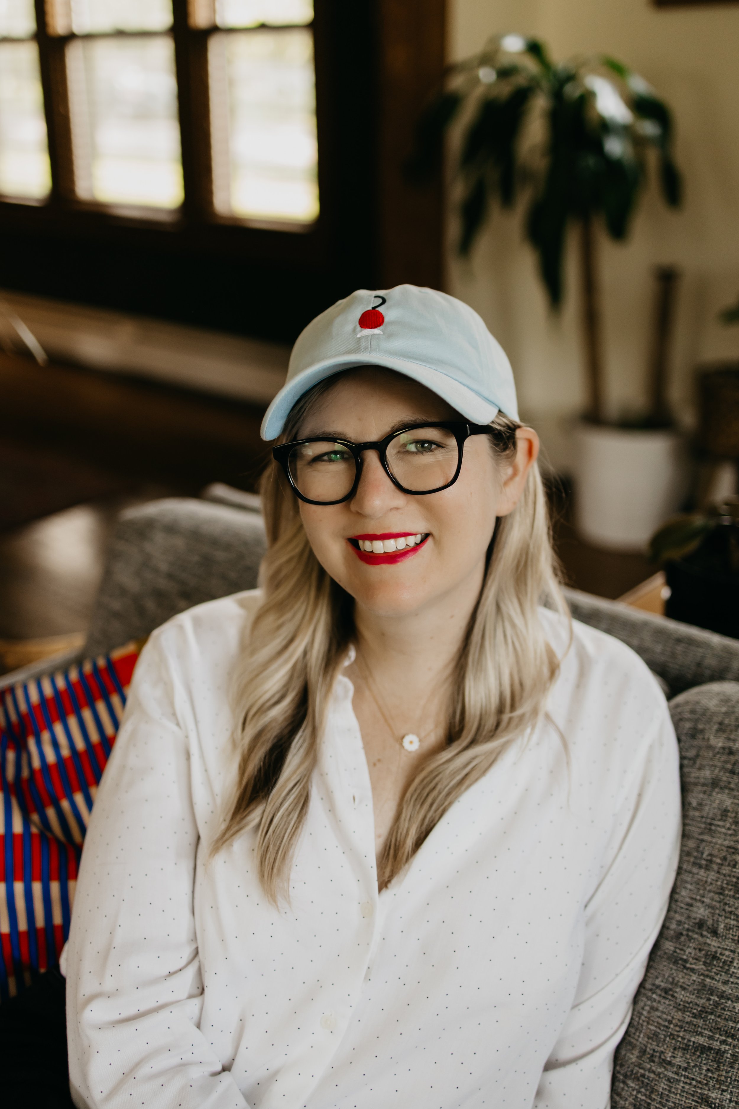 A woman with long blonde hair, black glasses, and red lipstick, wearing a white polka-dotted blouse and a white baseball cap with a small red and black design, smiling while sitting on a couch indoors.