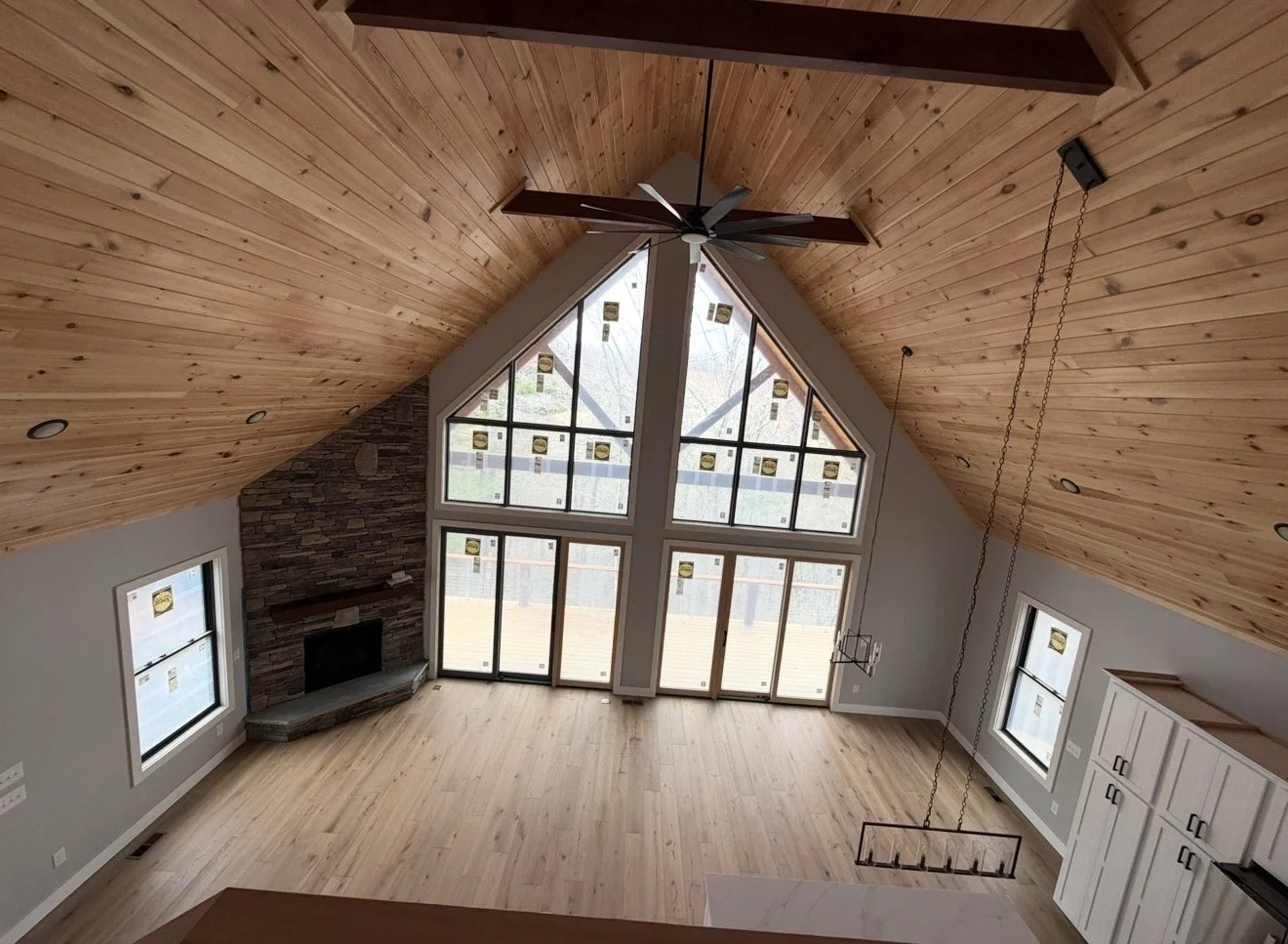 Interior view of a living room with a high vaulted wooden ceiling, large window with multiple panes, a stone fireplace, and a ceiling fan.