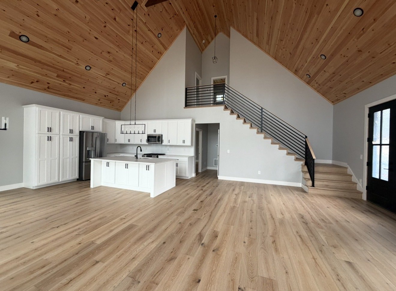 Open concept living room and kitchen with wood floors, white cabinets, stainless steel appliances, a kitchen island, a staircase with black metal railing, and a vaulted wooden ceiling. Antonio's Construction serving Ashe and Watauga County 