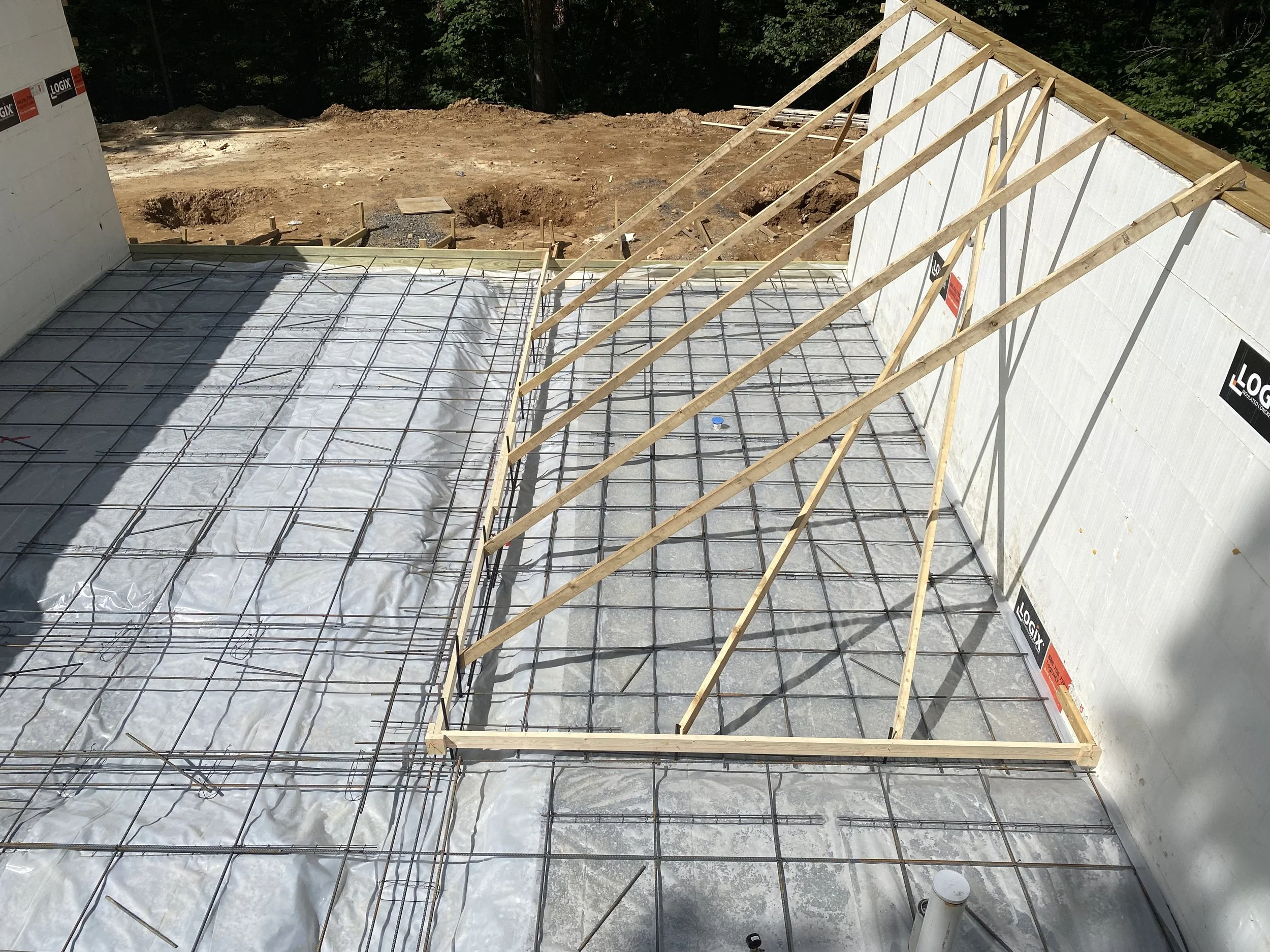 Construction site showing wooden framing and rebar for a building foundation, with insulated walls on two sides and outdoor dirt area in the background.