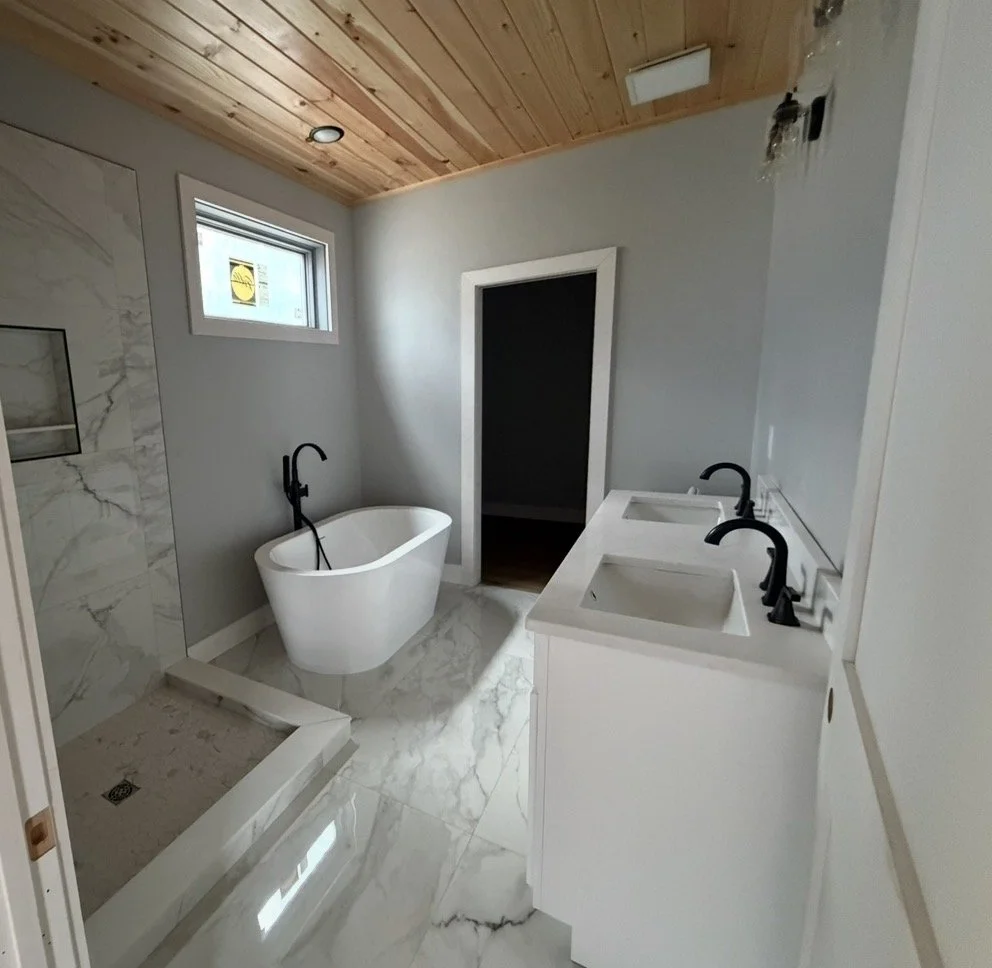A modern bathroom with a freestanding bathtub, dual sinks with black fixtures, a walk-in shower with marble tiles, a small window, and a wooden ceiling.