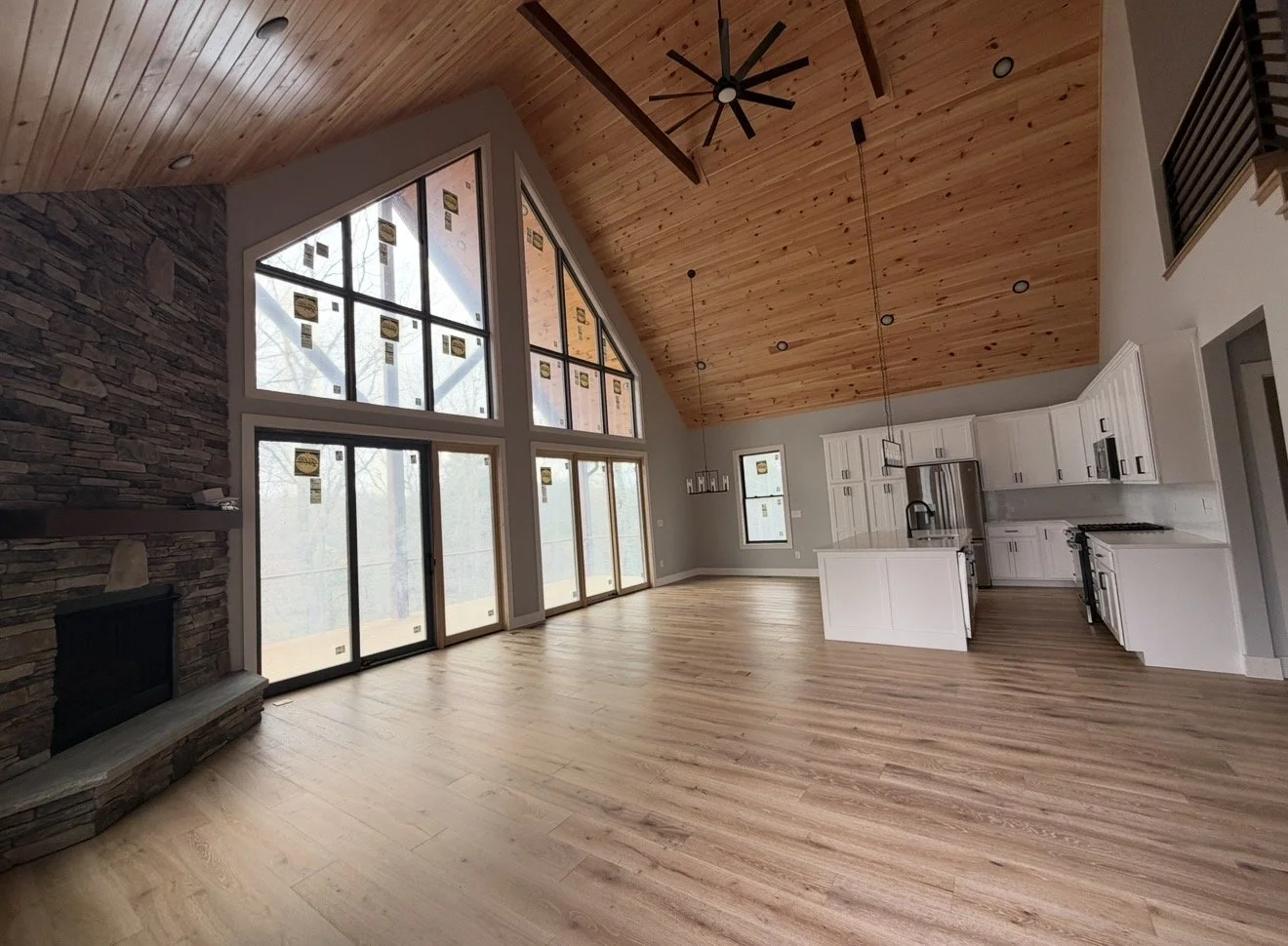 Open-concept living area with high vaulted wooden ceiling, large windows, stone fireplace, and white kitchen cabinets and island.