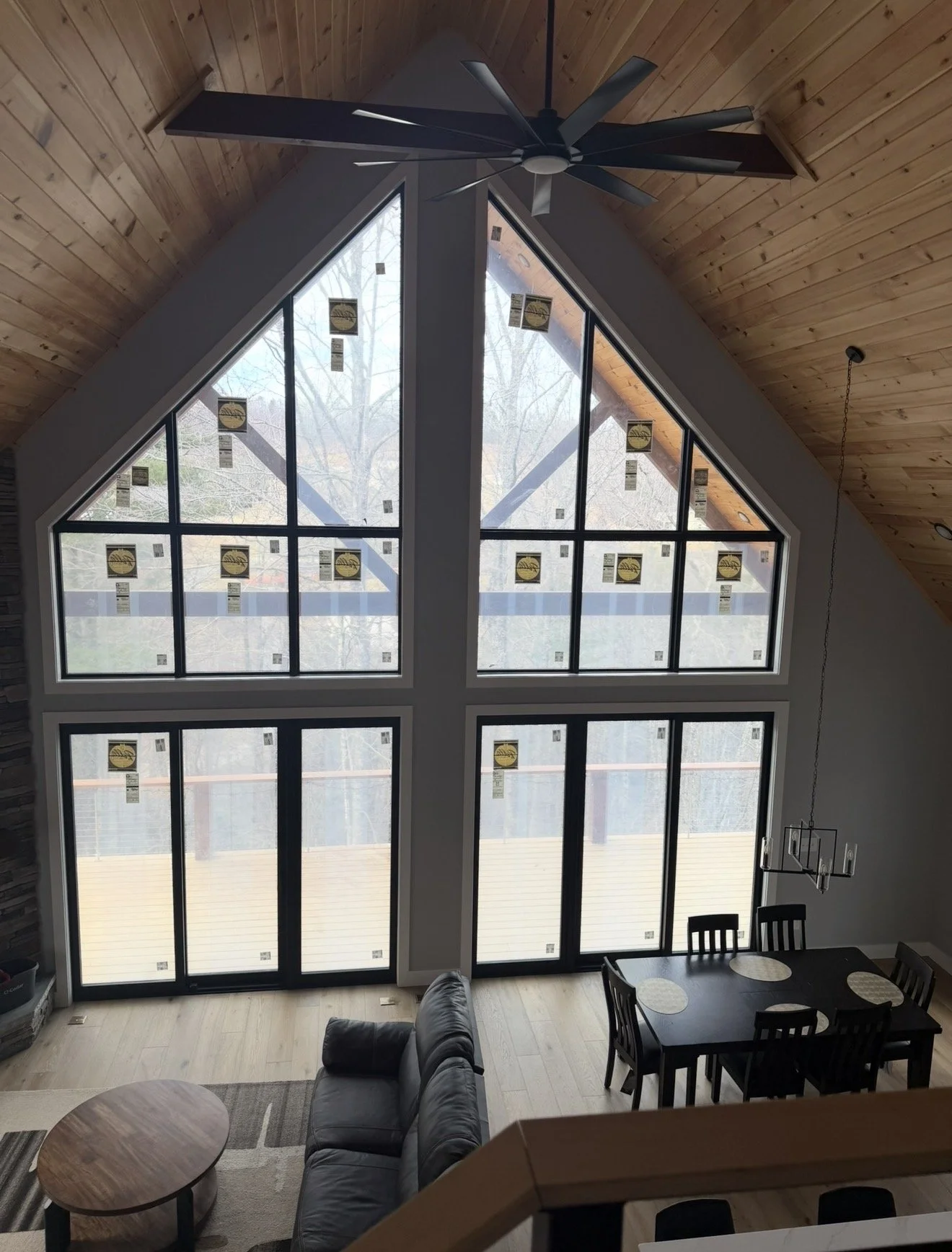 Interior of a house with large, triangular floor-to-ceiling windows, wooden vaulted ceiling, ceiling fan, and a dining area with a black table and chairs.