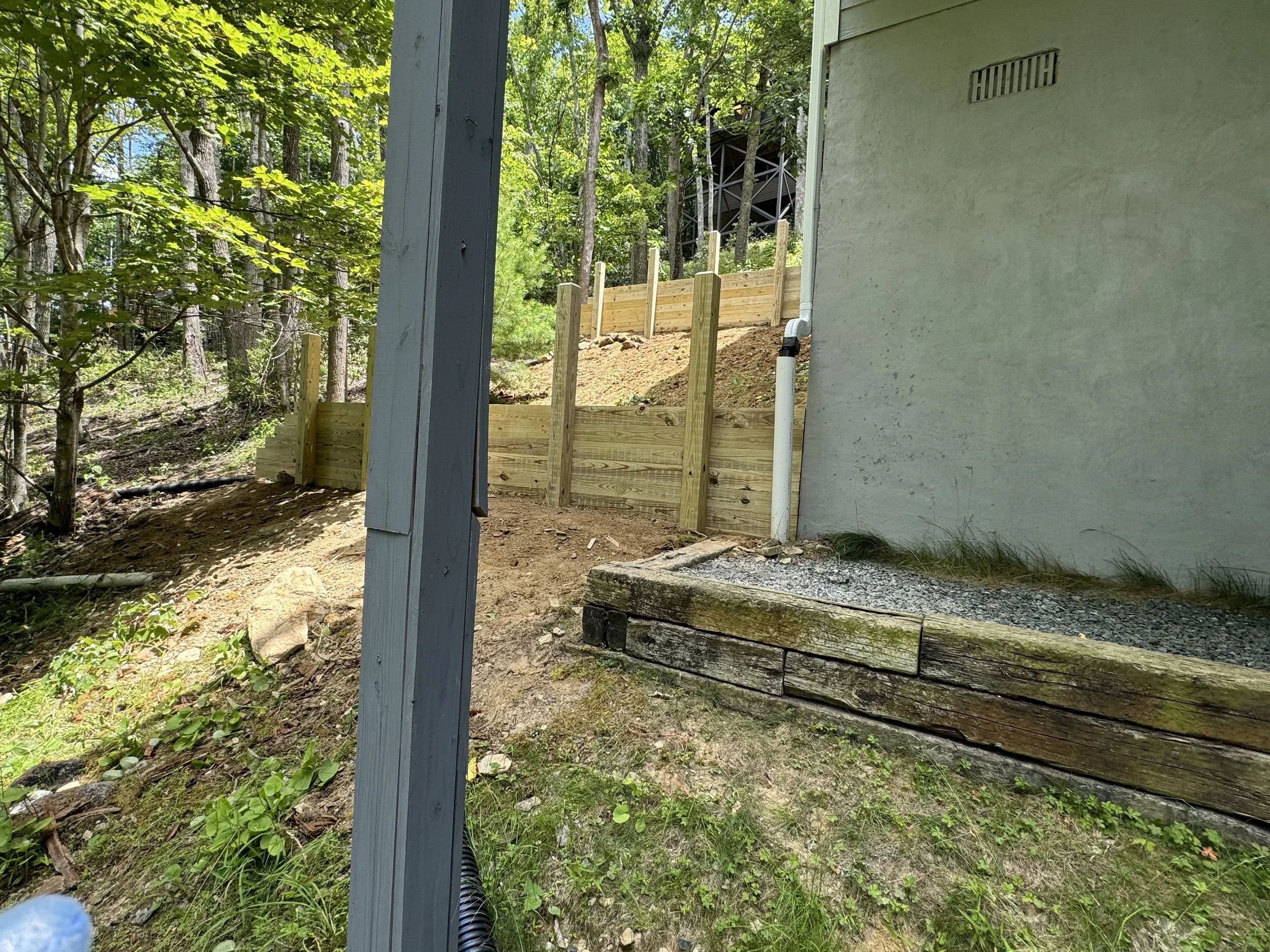 View of a backyard area with a new wooden retaining wall, a gray support post, and a white house wall. A drain pipe extends from the house, and the ground is uneven with soil, grass, and a gravel section. Trees and wooded area are visible in the back
