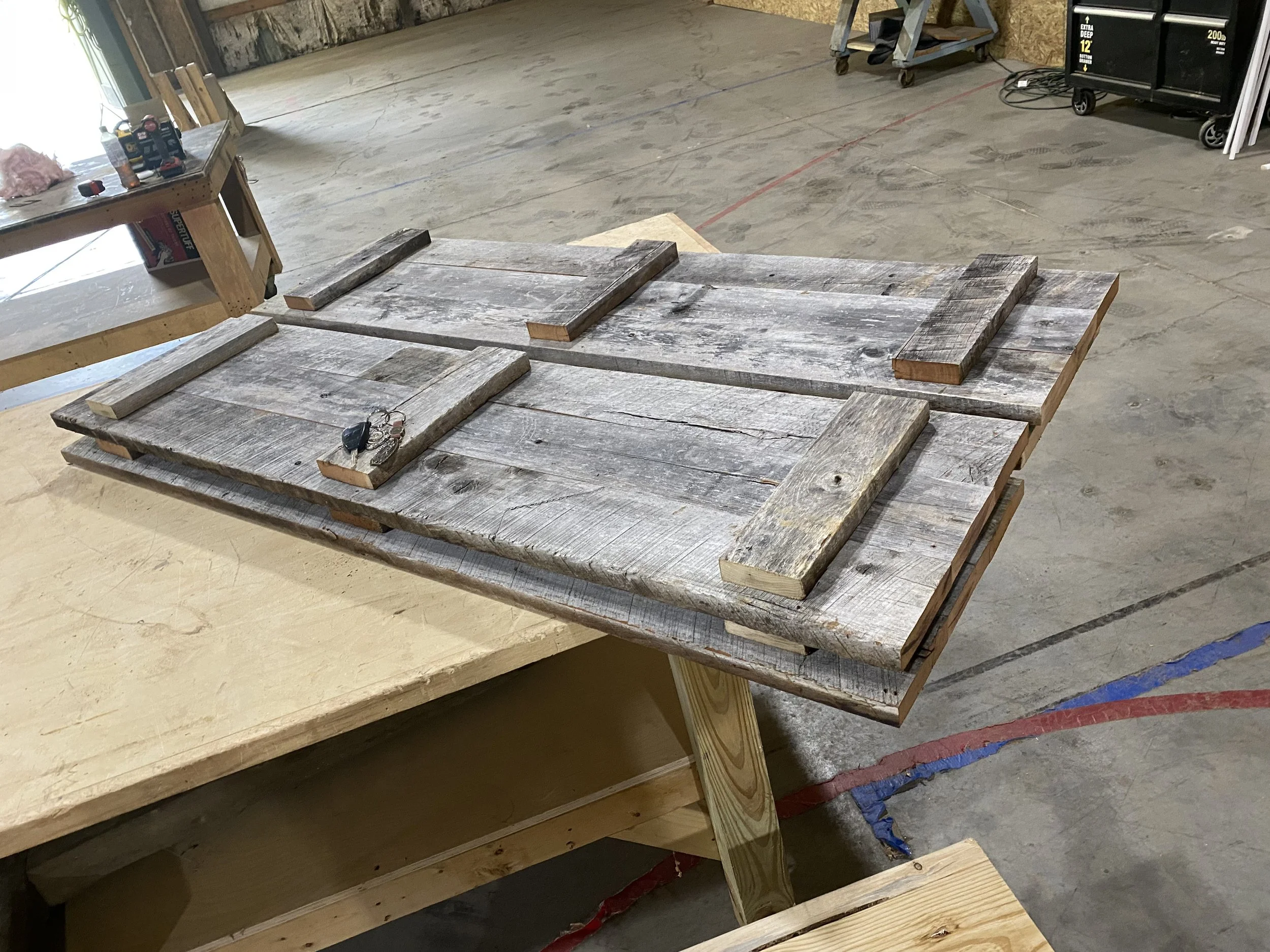 Wooden pallets and planks arranged on a workbench, in a workshop with concrete floor, tools, and equipment in the background.