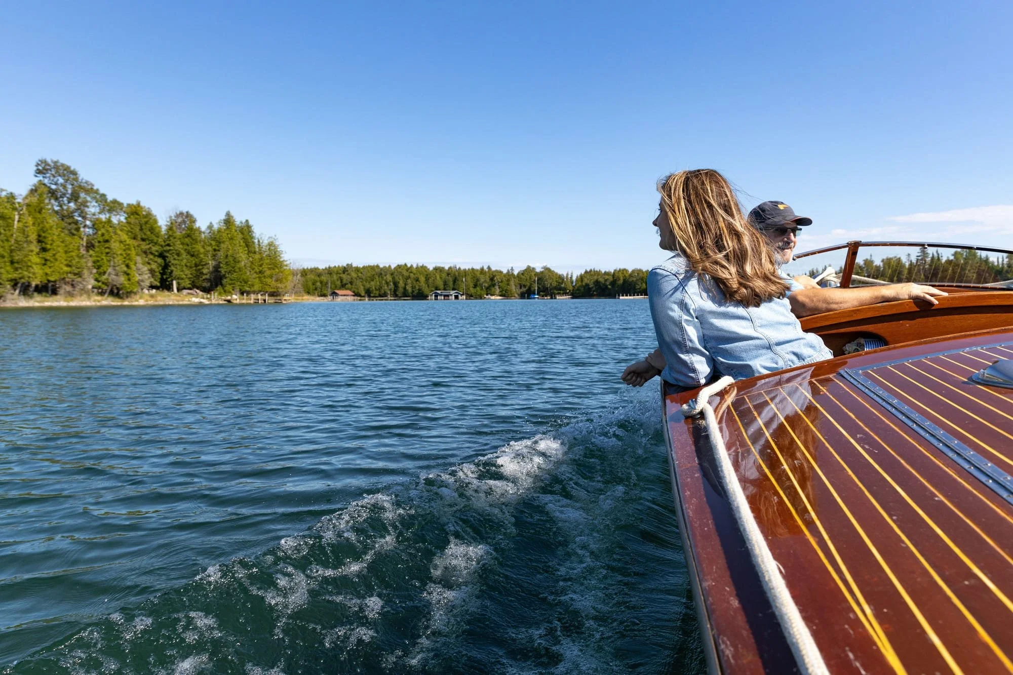 A woman with long hair and a man wearing a cap and sunglasses enjoy a boat ride on a lake with trees and small cabins in the background under a clear blue sky.
