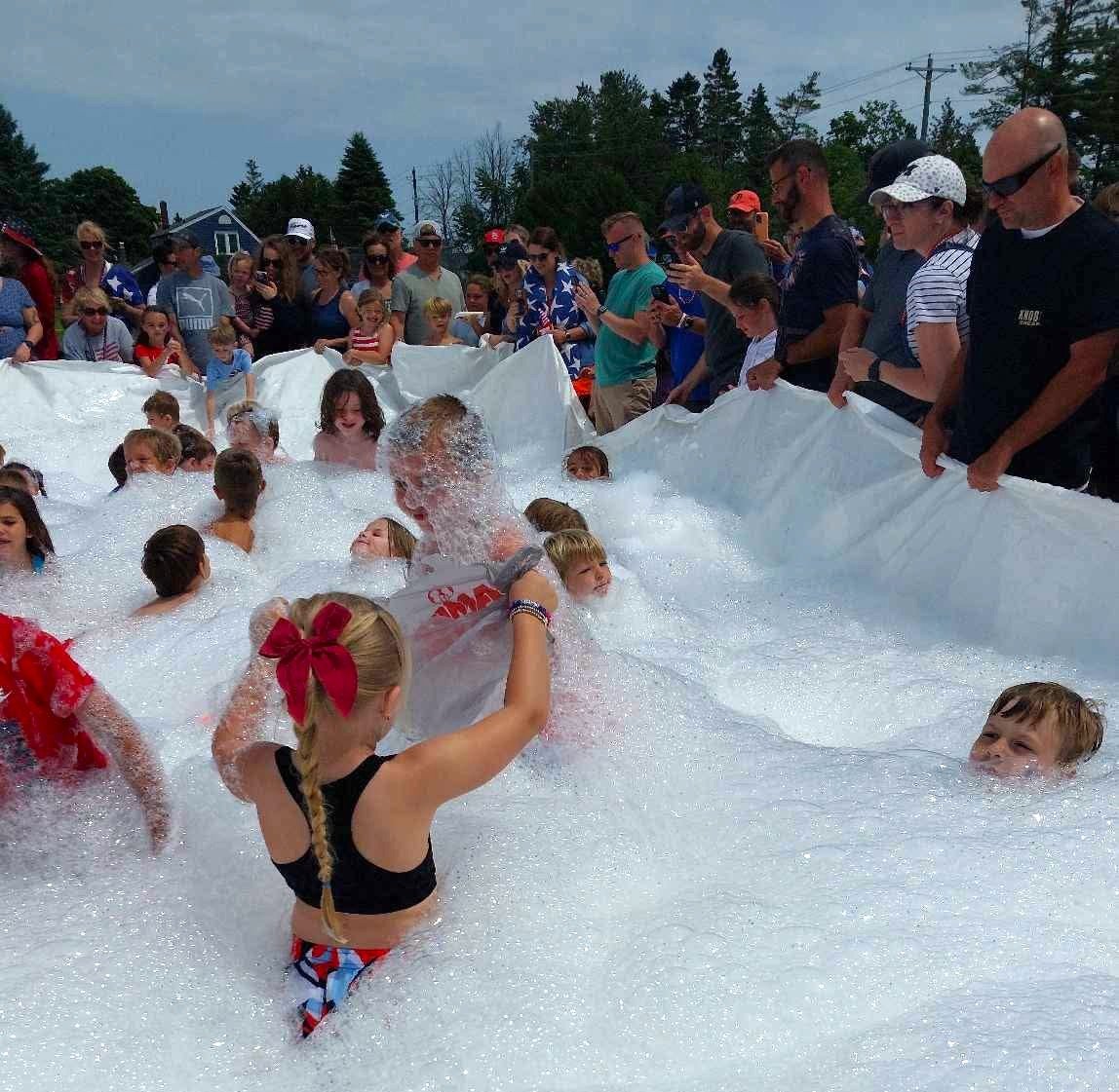 Children and adults enjoying a foam-filled outdoor event, with some children playing and adults watching.