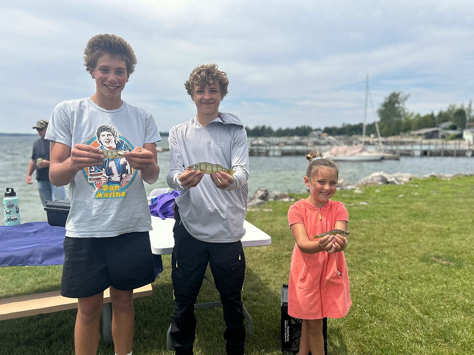 Three children posing outdoors by a body of water, holding small fish they caught. Two boys and a girl smiling, with boats and docks in the background.