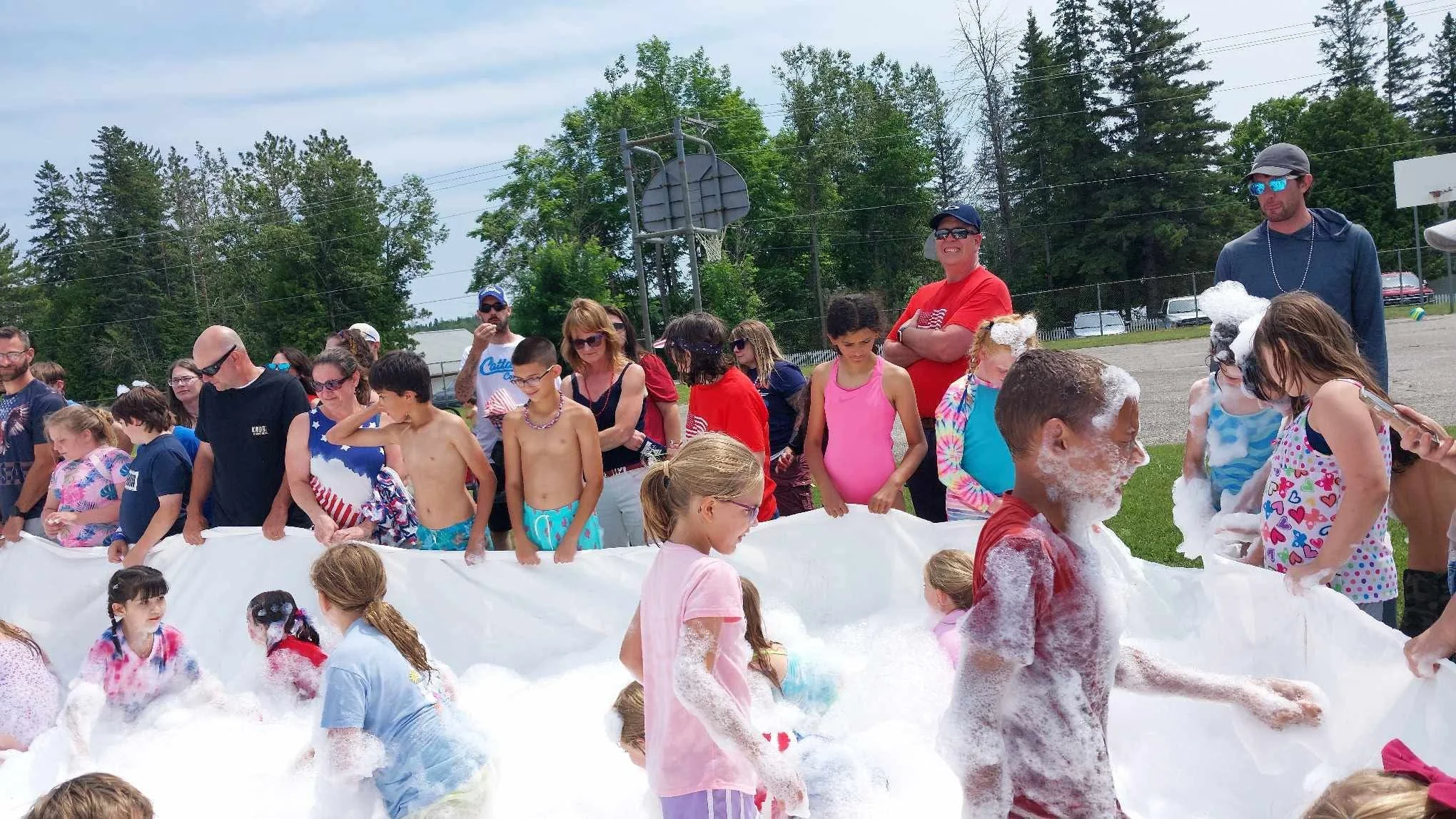 Children and adults participating in a foam-filled outdoor event on a sunny day, with trees and a basketball hoop in the background.