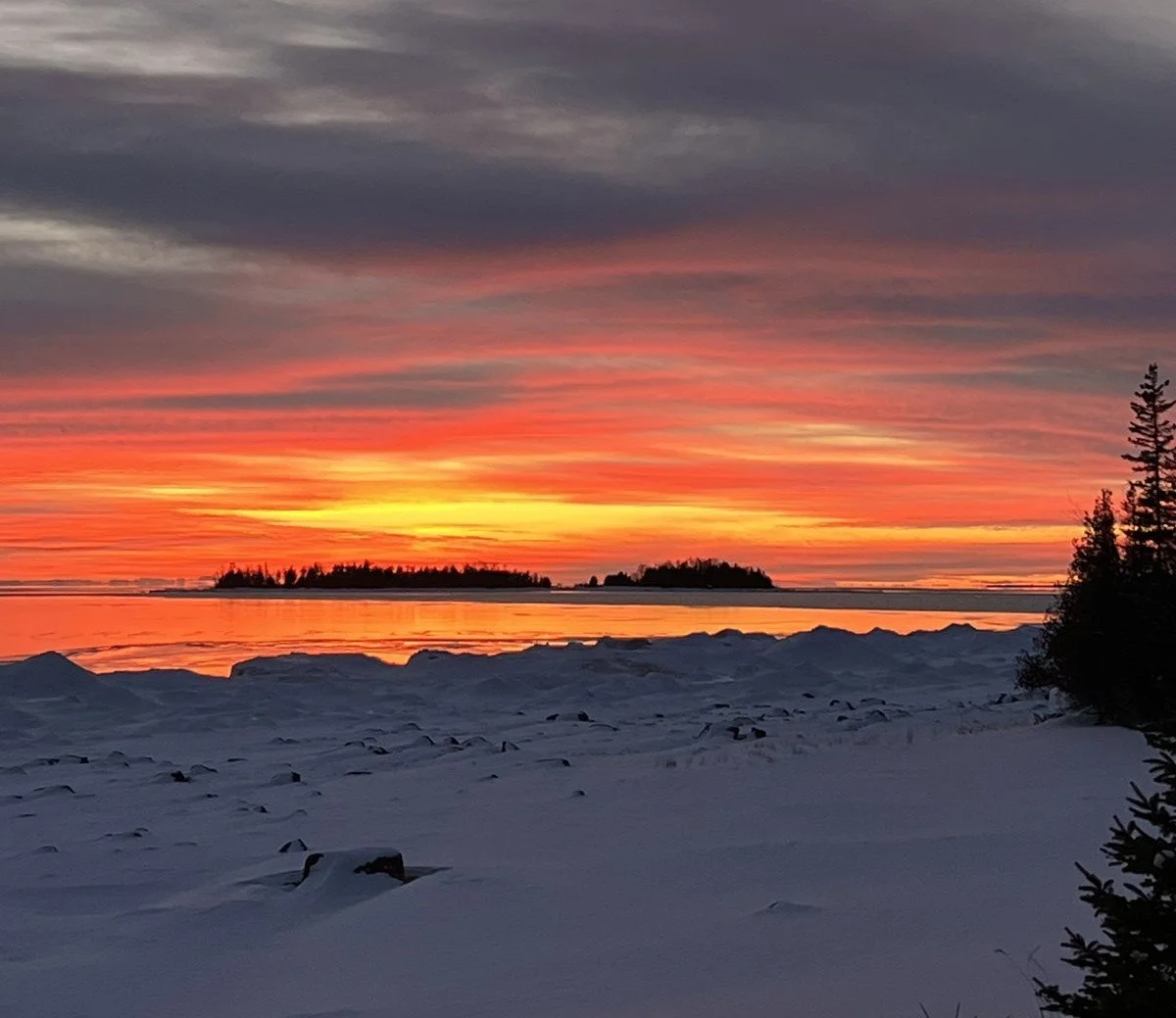 Snow-covered shoreline with a calm body of water and small islands in the distance, vibrant sunset sky with orange, pink, and purple hues, and dark silhouettes of trees on the right.