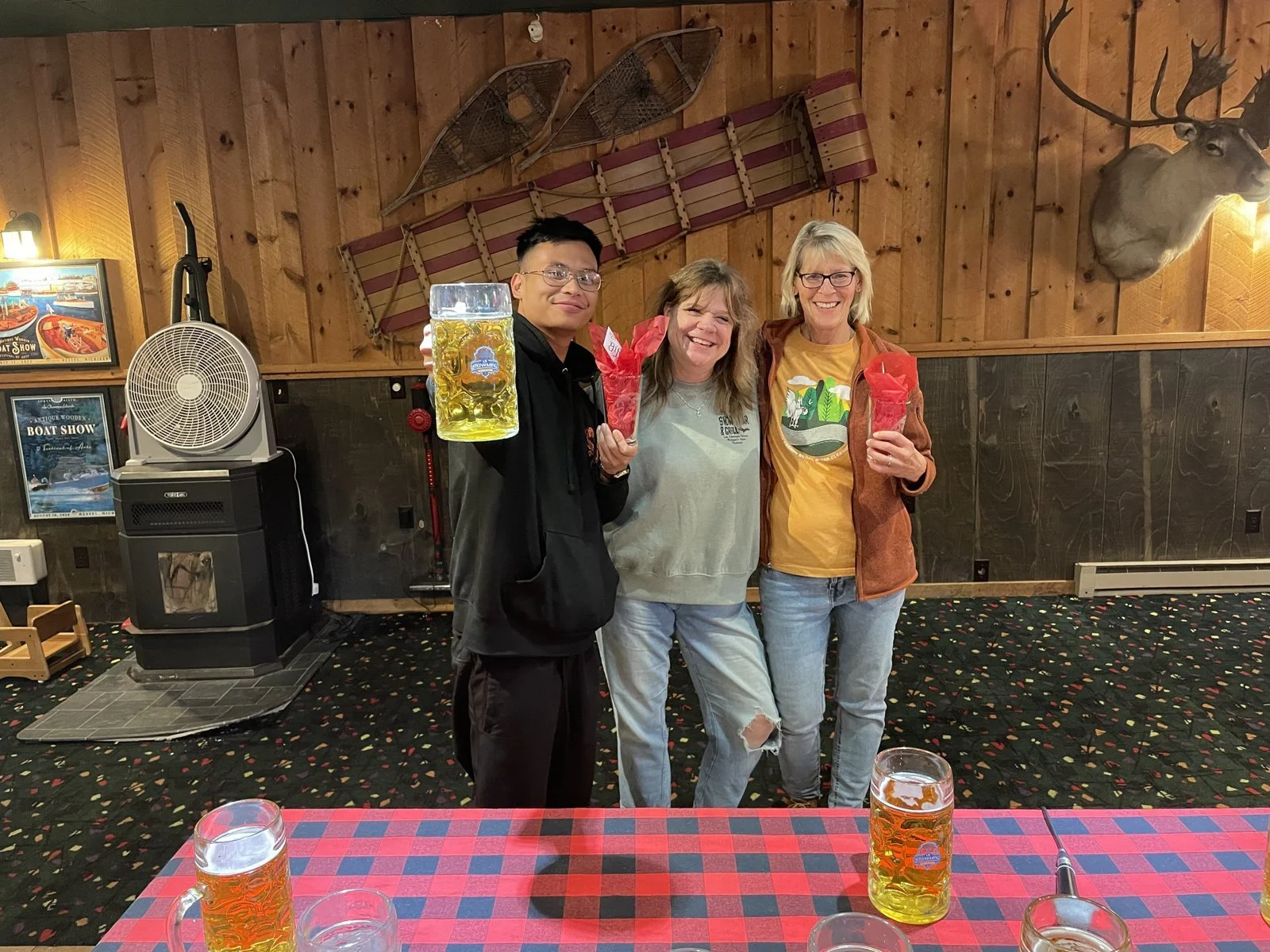 Three people standing together indoors, smiling, holding drinks and gift bags. The background features wooden walls, vintage sleds, a mounted moose head, and a fan. There are glasses of beer on a red checkered table in the foreground.