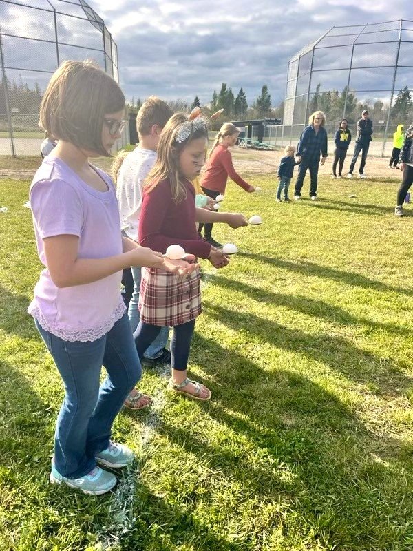 Children and adults participating in an egg relay race on a baseball field, holding eggs in spoons.
