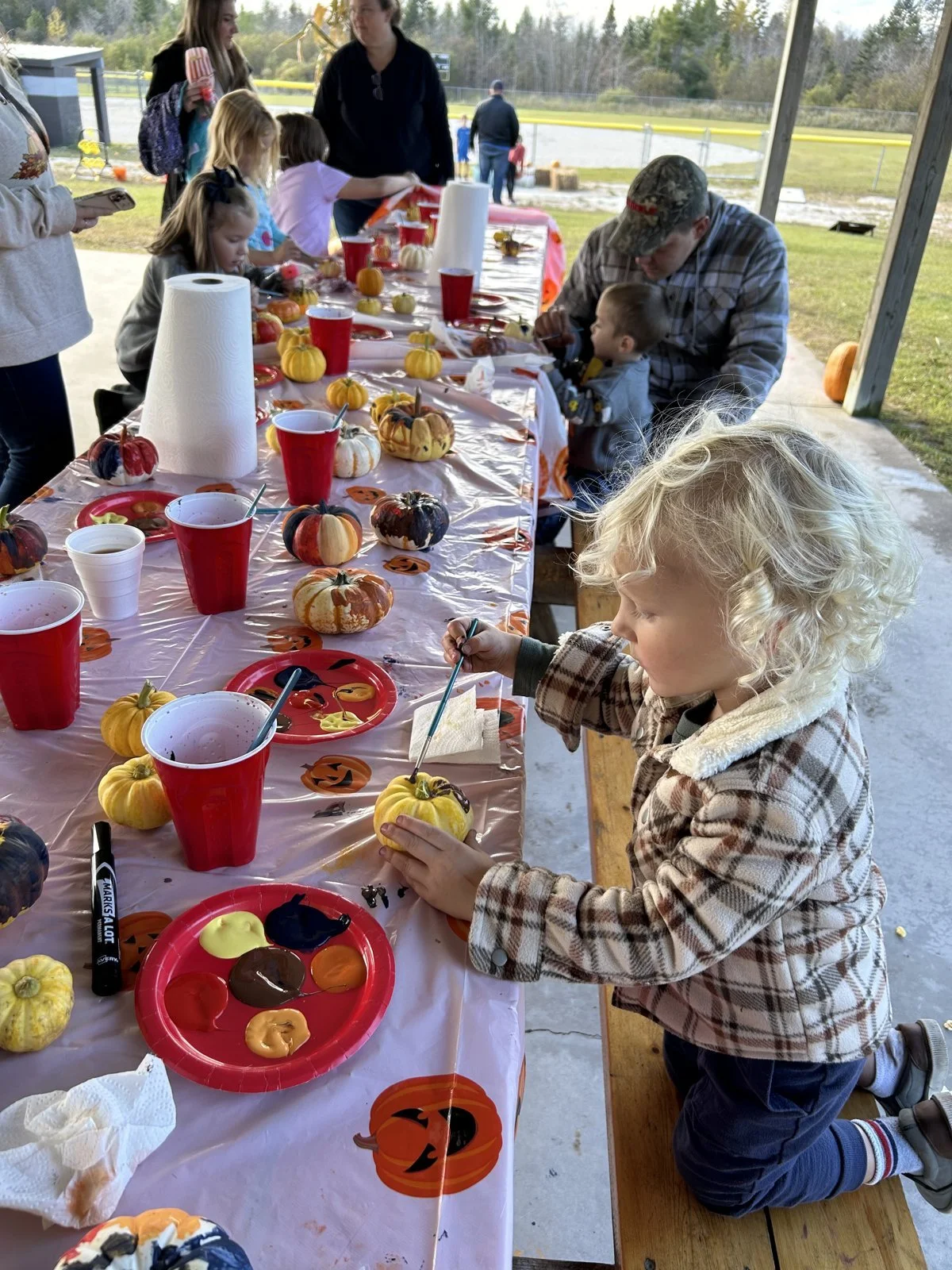 Children and adults at a fall-themed pumpkin decorating party outdoors, with pumpkins painted in various colors and patterns, and paint palettes on a picnic table.