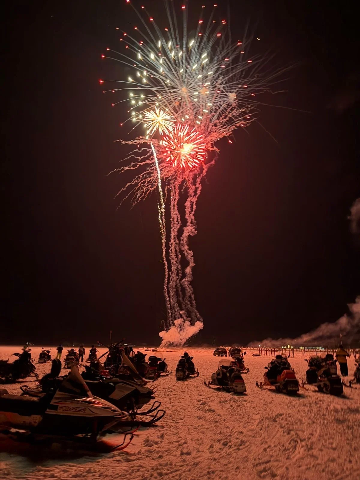 Nighttime fireworks display above a snow-covered area with parked snowmobiles and motorcycles, and a crowd watching.