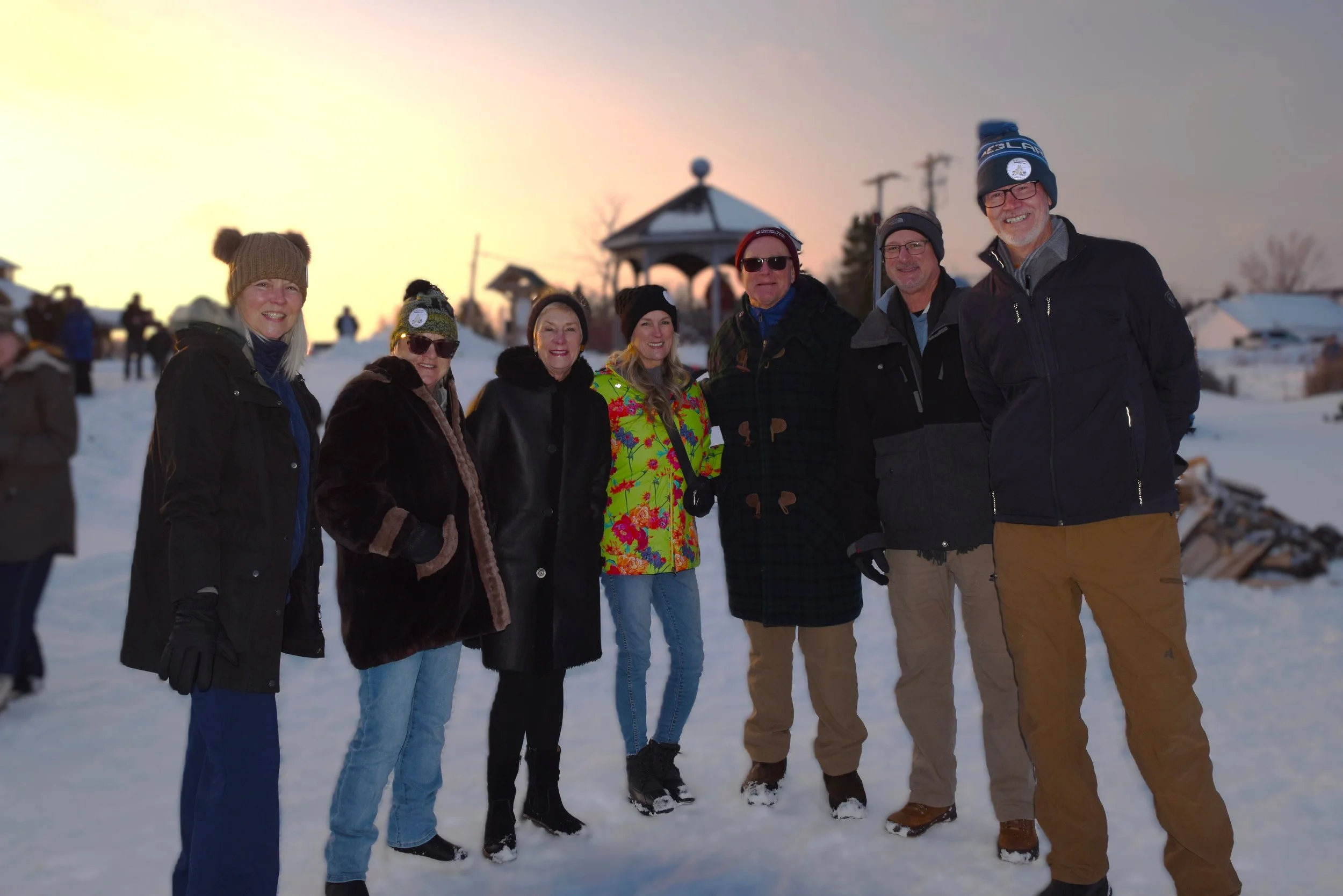 A group of seven people standing outside in the snow during sunset, dressed in winter clothing, smiling at the camera.