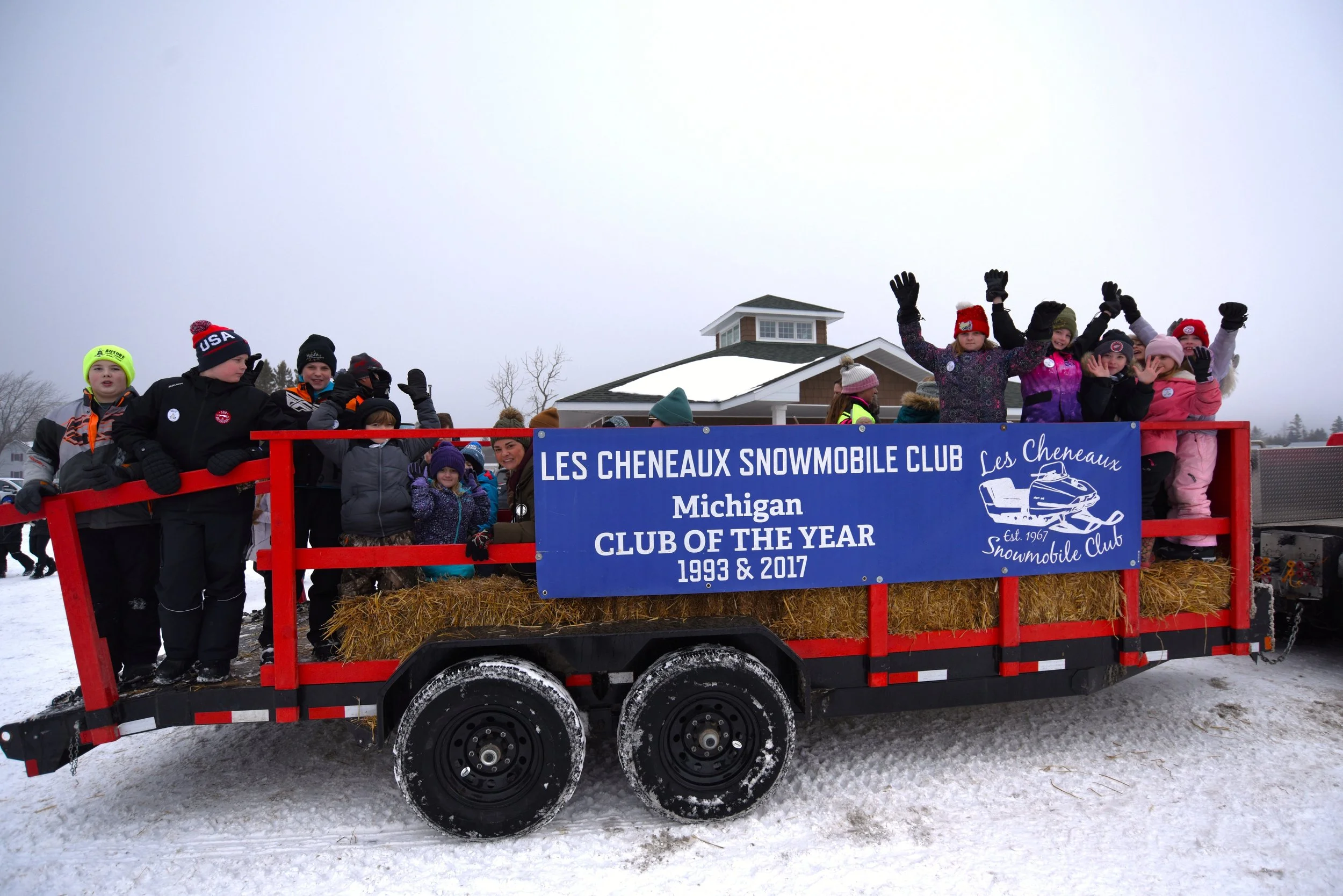 Group of children and adults on a snowmobile trailer with a banner that reads "Les Cheneaux Snowmobile Club, Michigan, Club of the Year 1993 & 2017," all dressed warmly in winter clothing, waving and smiling in a snowy outdoor setting.