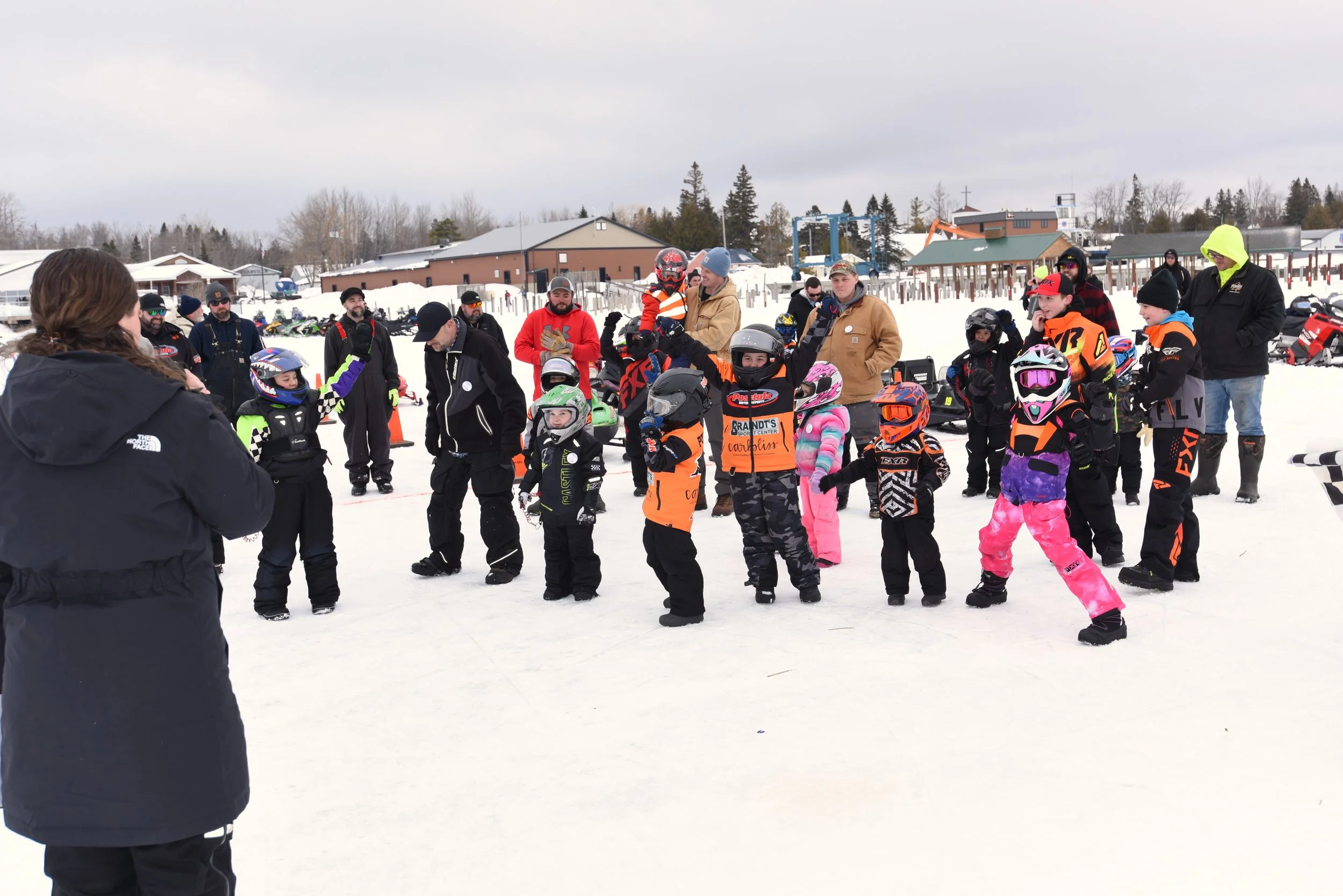 A group of children and adults gathered on snow, some children wearing colorful winter gear and helmets, with sleds and snowmobiles in the background, at a snowy outdoor location.