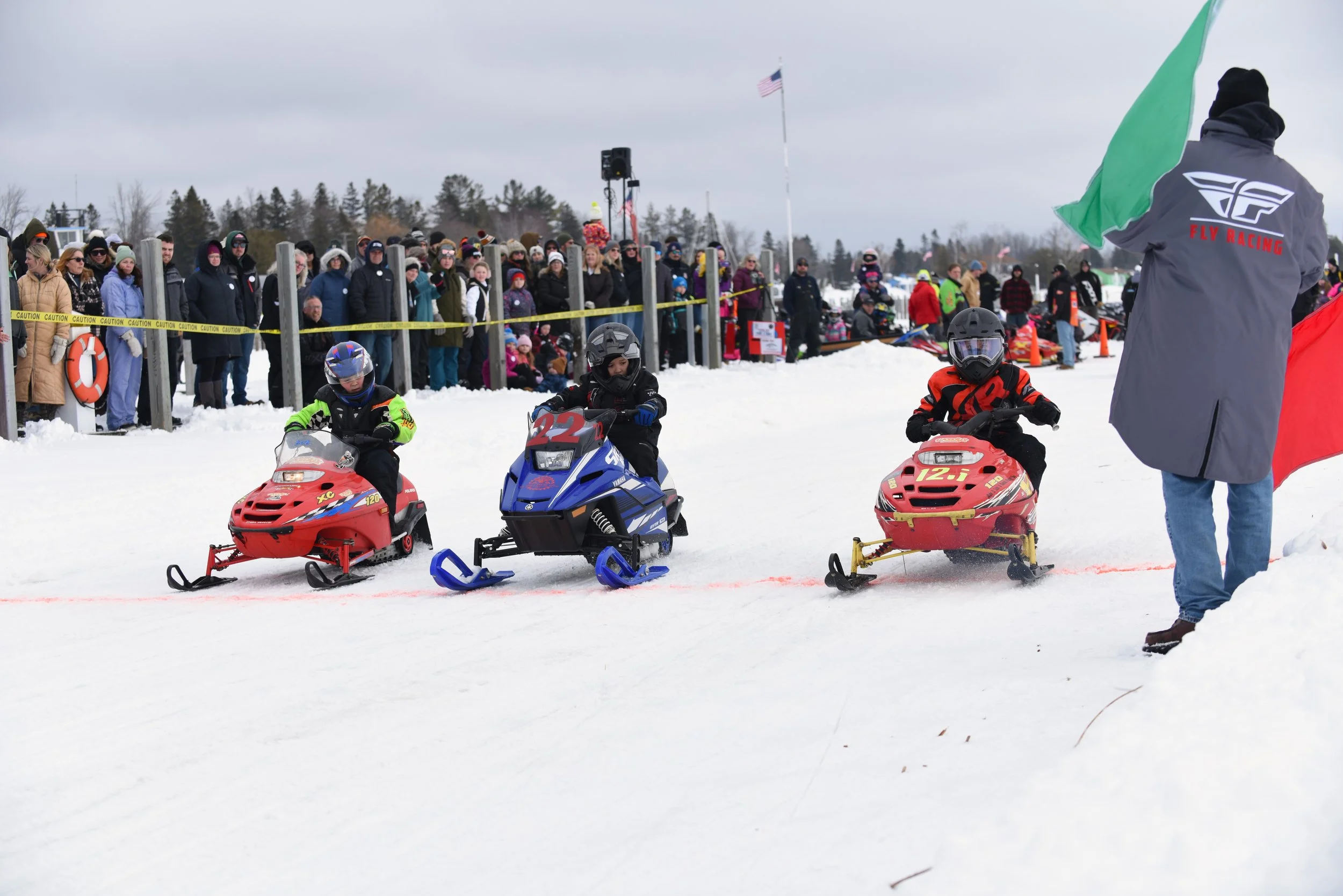 Children riding snowmobiles during a winter event, with spectators and a flag in the background.