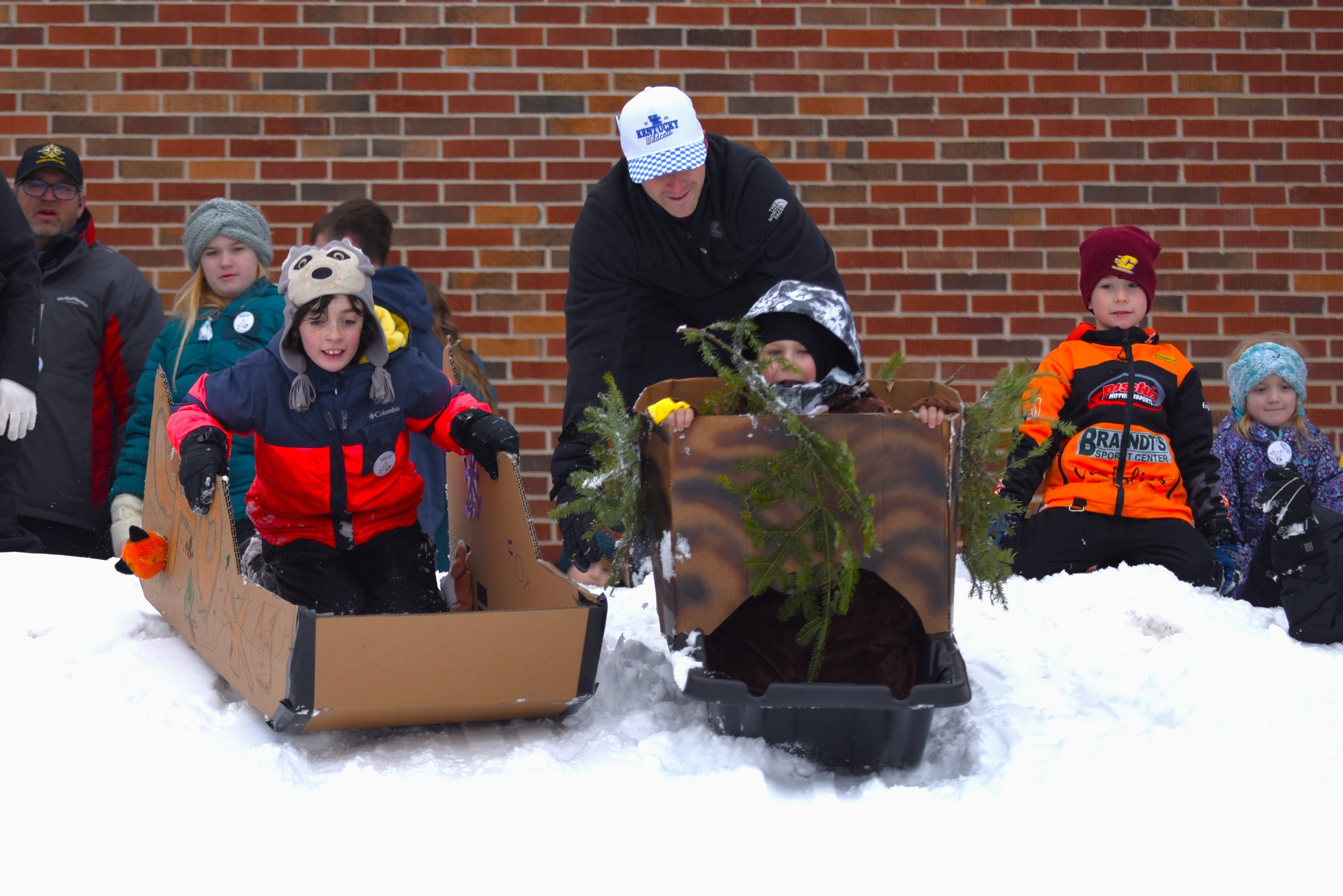 Children and adults in winter clothing standing behind a sled filled with pine branches and decorative greenery, as part of an outdoor winter event against a brick wall.