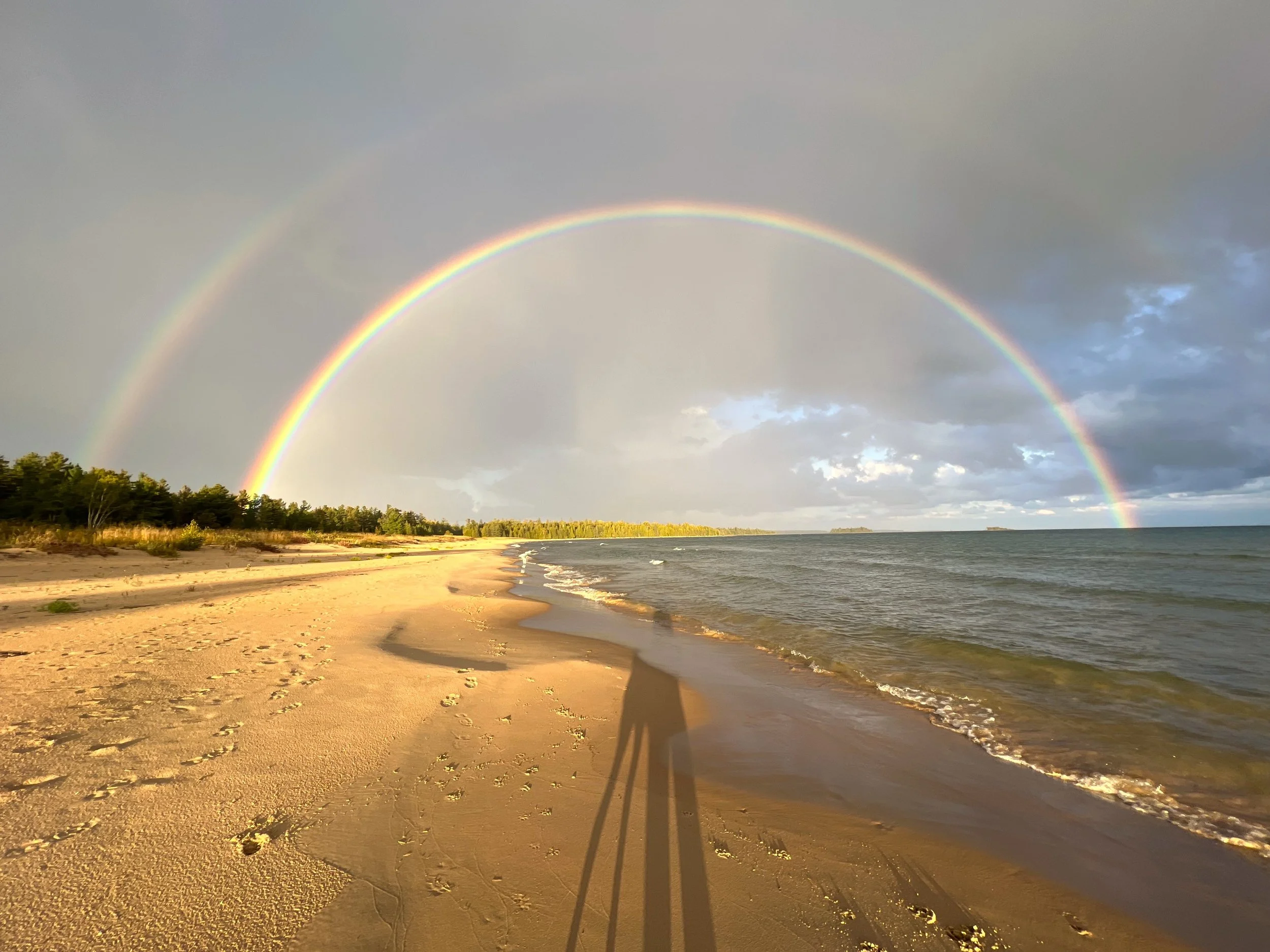 A beach with sand and footprints, calm ocean waves, dense trees in the distance, a double rainbow in a cloudy sky, and a shadow of a person or object on the sand.