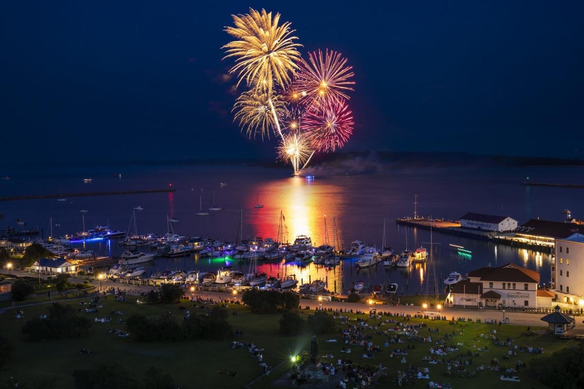 Fireworks exploding over a harbor at night with boats and people watching from the shore.