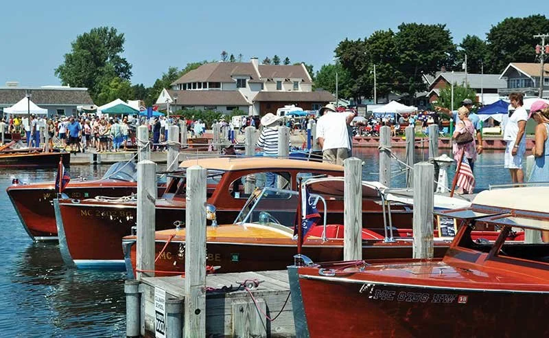People walking along a marina with wooden boats docked, near a waterfront area with houses and tents.