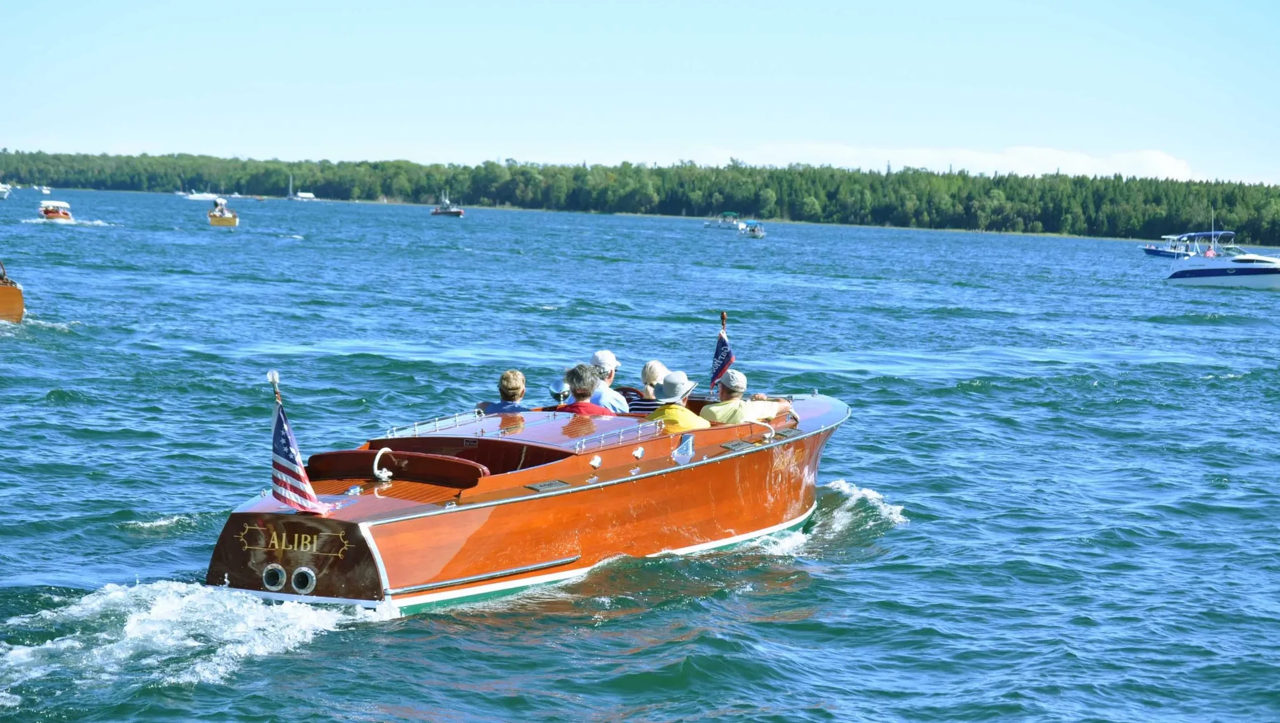 A wooden boat named 'Alibi' with several people on board sailing on a lake or river, surrounded by boats and lush green trees in the background.
