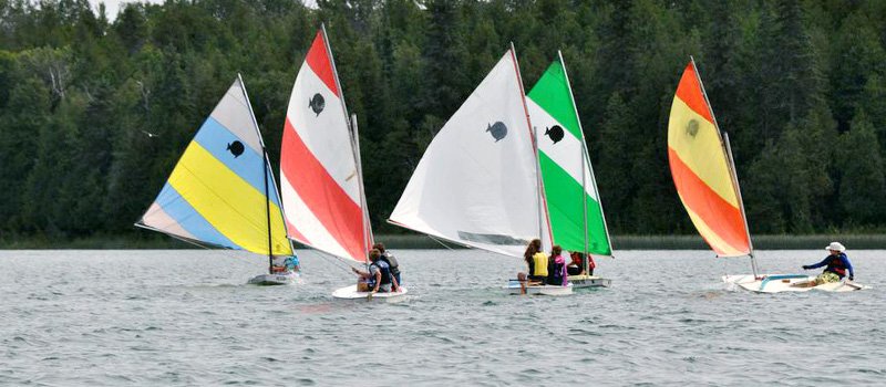 Four small sailboats with colorful sails, each with a person on board, sailing on a lake with a forested shoreline in the background.