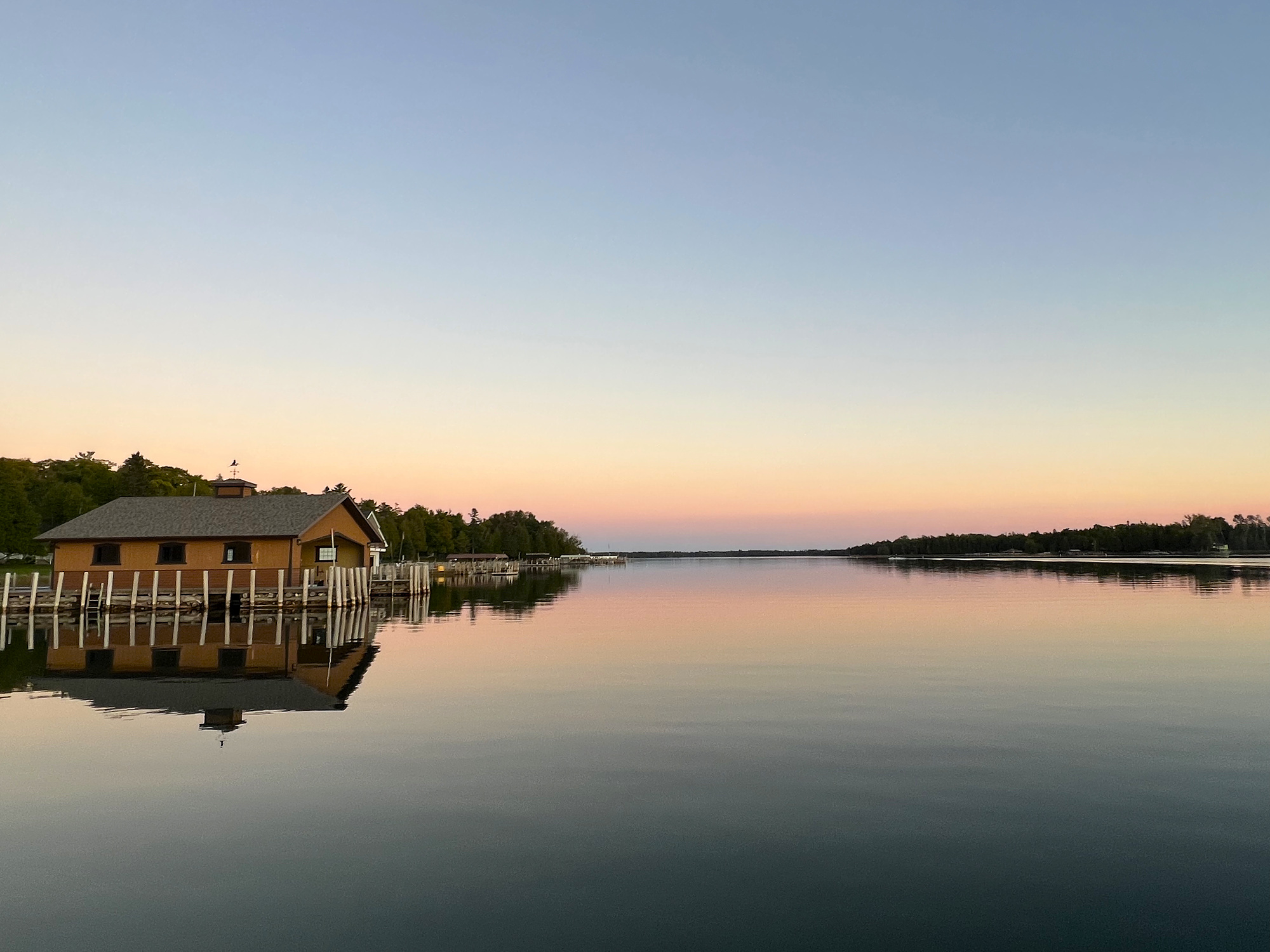 A peaceful scene of a house on stilts by the water at sunset, with a clear sky and calm water reflecting the sky and house.