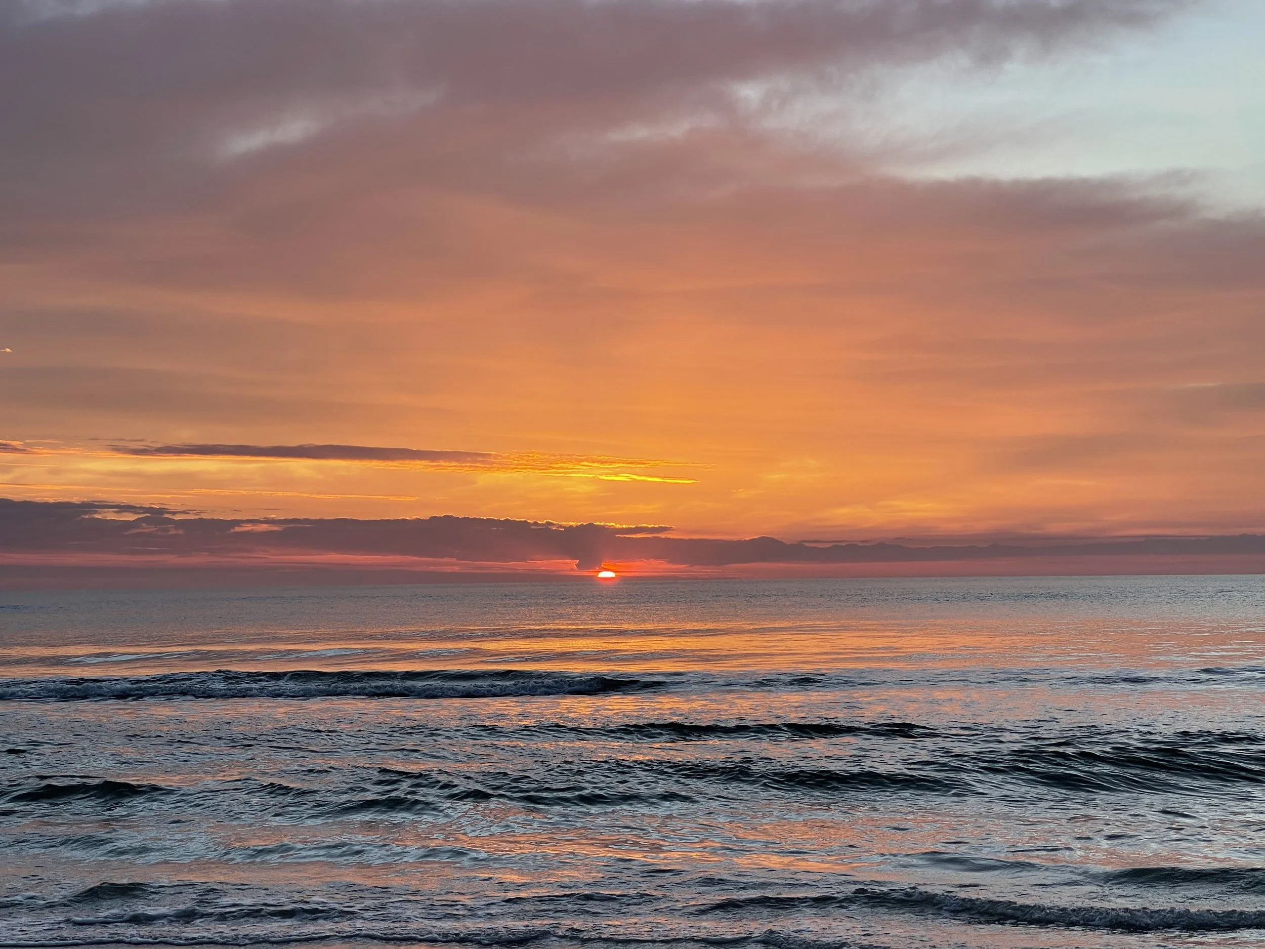 Sunset over the ocean with clouds and a distant horizon, colors ranging from orange and yellow to purple and pink, with gentle waves in the foreground.