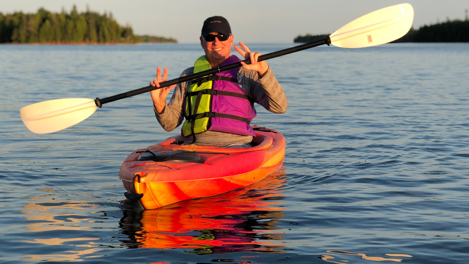 A smiling man in sunglasses paddling a red and orange kayak on a calm lake, wearing a purple and yellow life jacket during sunset.
