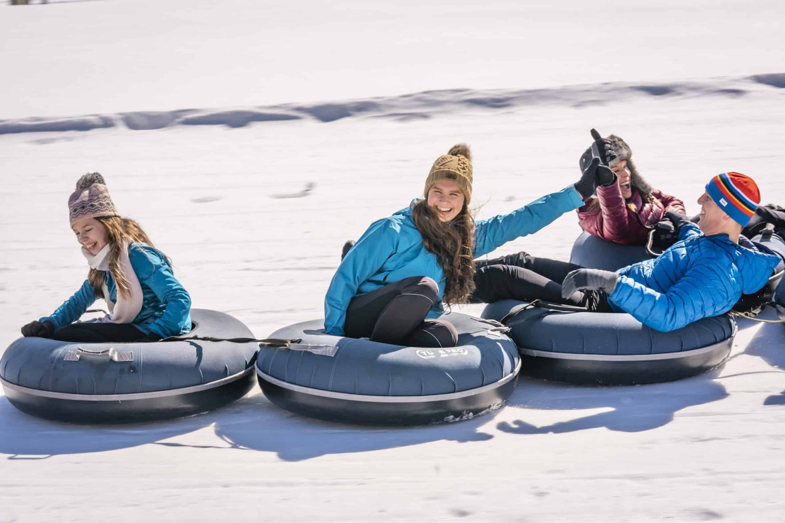 Group of four people riding inflatable snow tubes in the snow, smiling and enjoying winter outdoor activity.